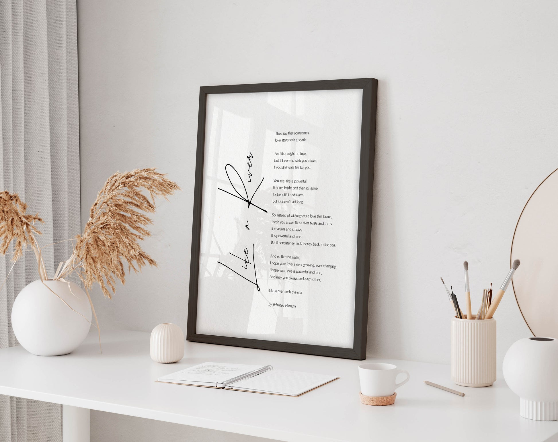 a framed poem titled "River" displayed on a white desk, accompanied by a notebook, a cup, and a vase with dried flowers.