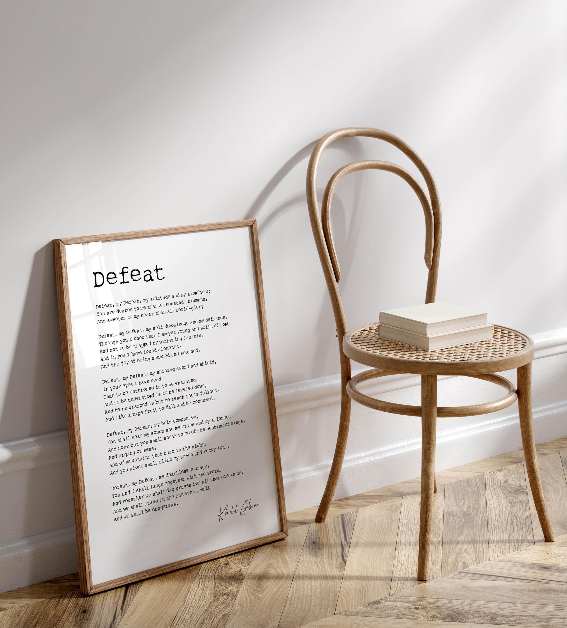 a framed poem titled " Defeat" leaning against a wall, with a wooden chair and a stack of books placed next to it on a wooden floor.