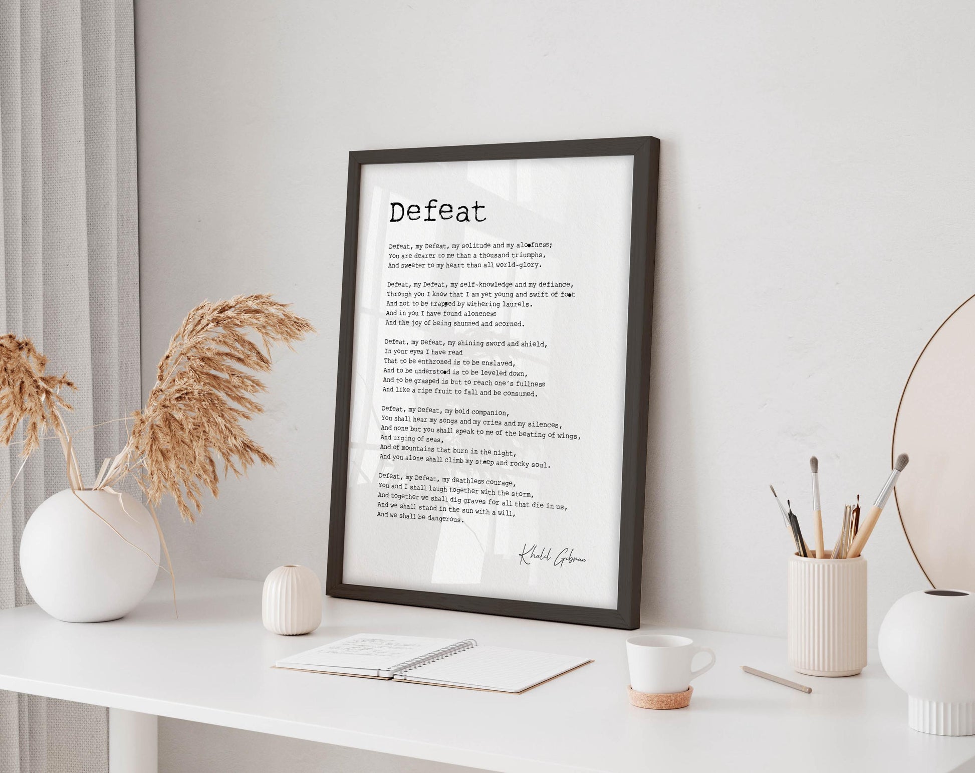a framed poem titled "Defeat" displayed on a white wall, with a notebook, pen, and other office supplies arranged in front of it.