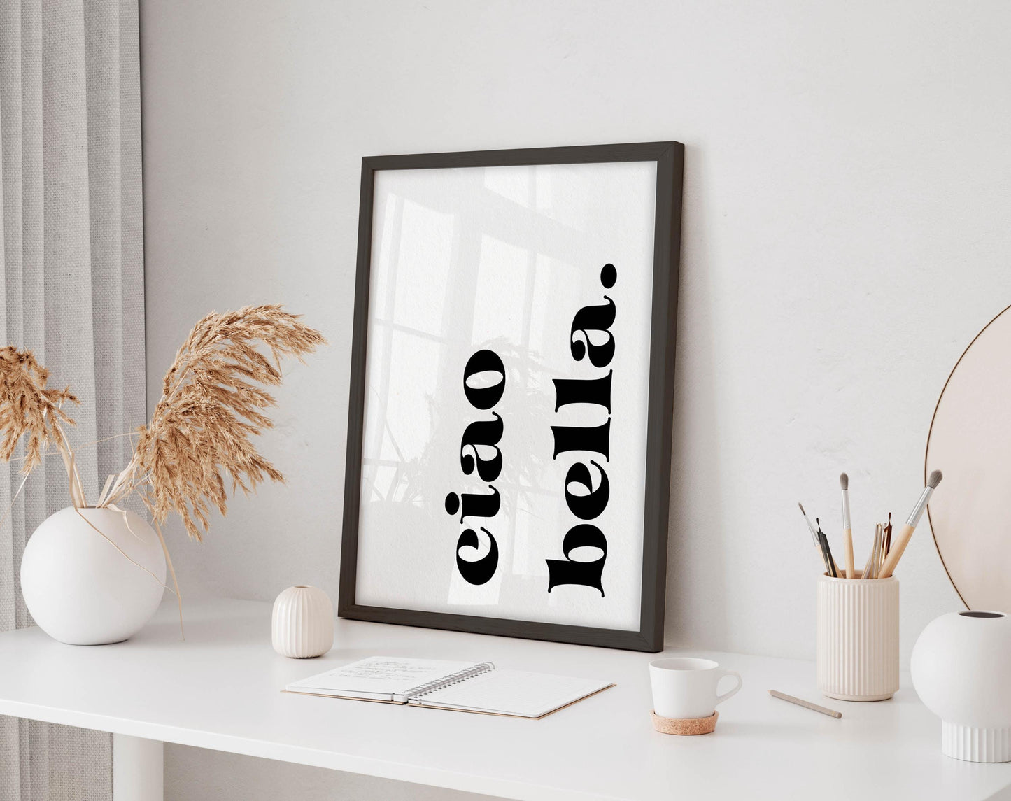 a white desk with a framed poster, a notebook, a cup, and a vase with dried flowers.
