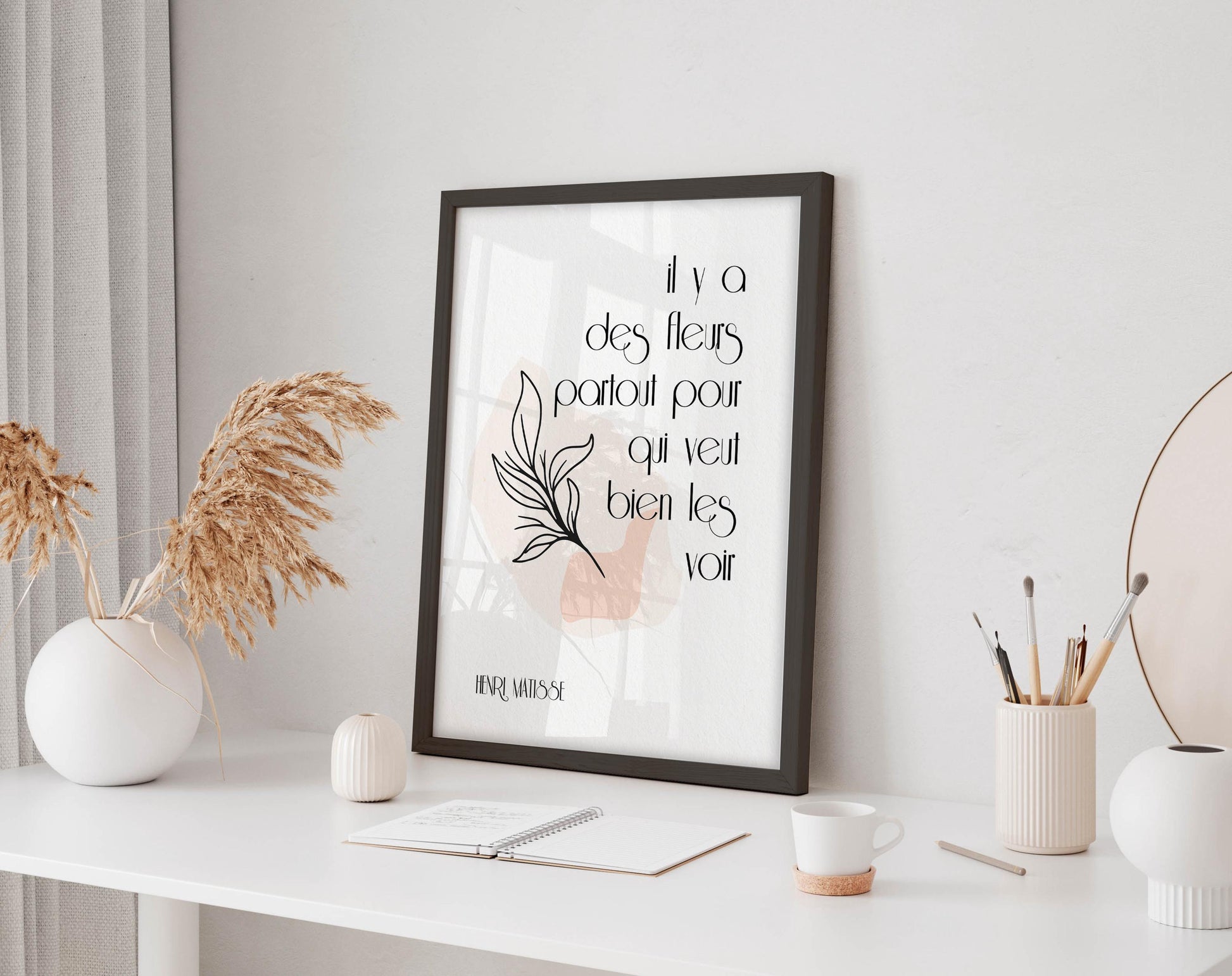 a framed quote on a white desk, surrounded by various objects such as a notebook, a cup, and a vase with dried flowers.