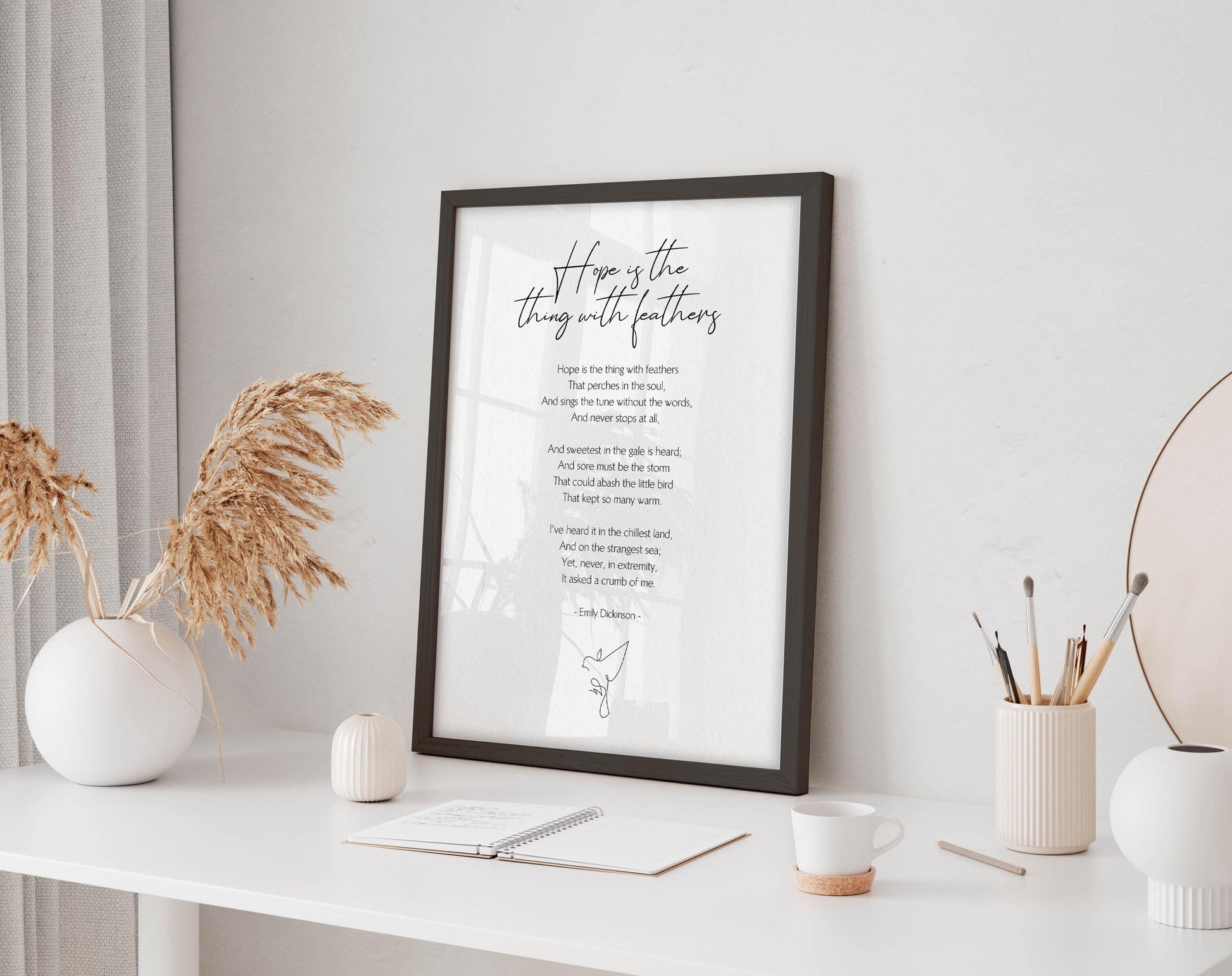 a framed poem titled "It's the thing with feathers" displayed on a white desk, accompanied by a notebook, a cup, and a vase with dried flowers.