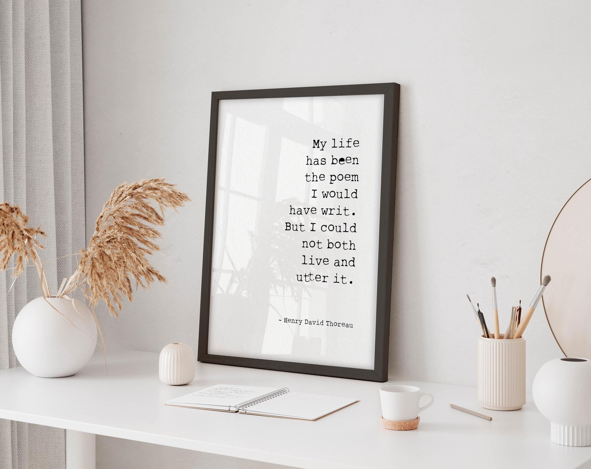 a framed quote on a white desk, surrounded by various objects such as a notebook, a cup, and a vase with dried flowers.