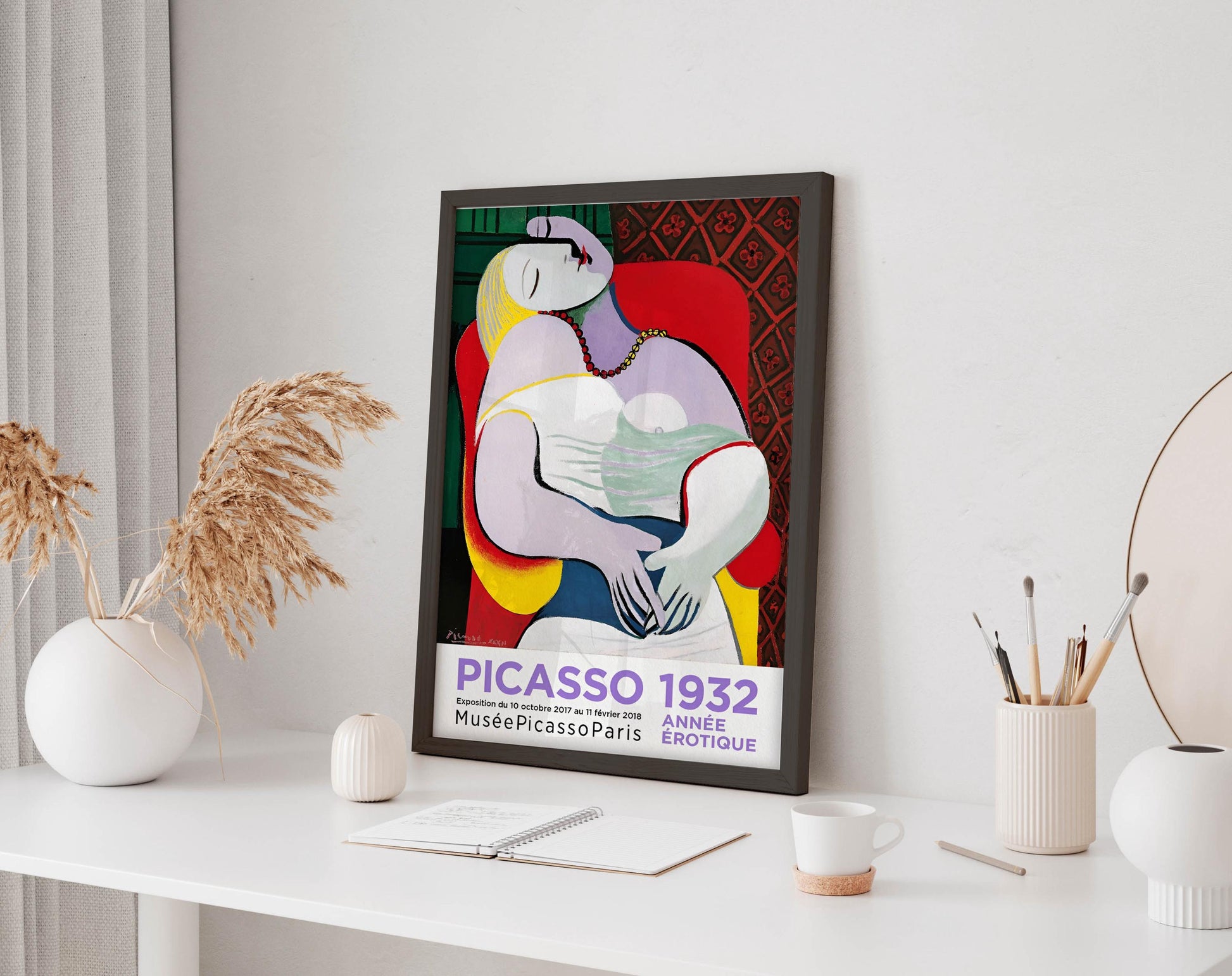 a framed poster of the cubist painting "PICasso 1922" displayed on a white desk, surrounded by various objects such as a notebook, a cup, and a vase.