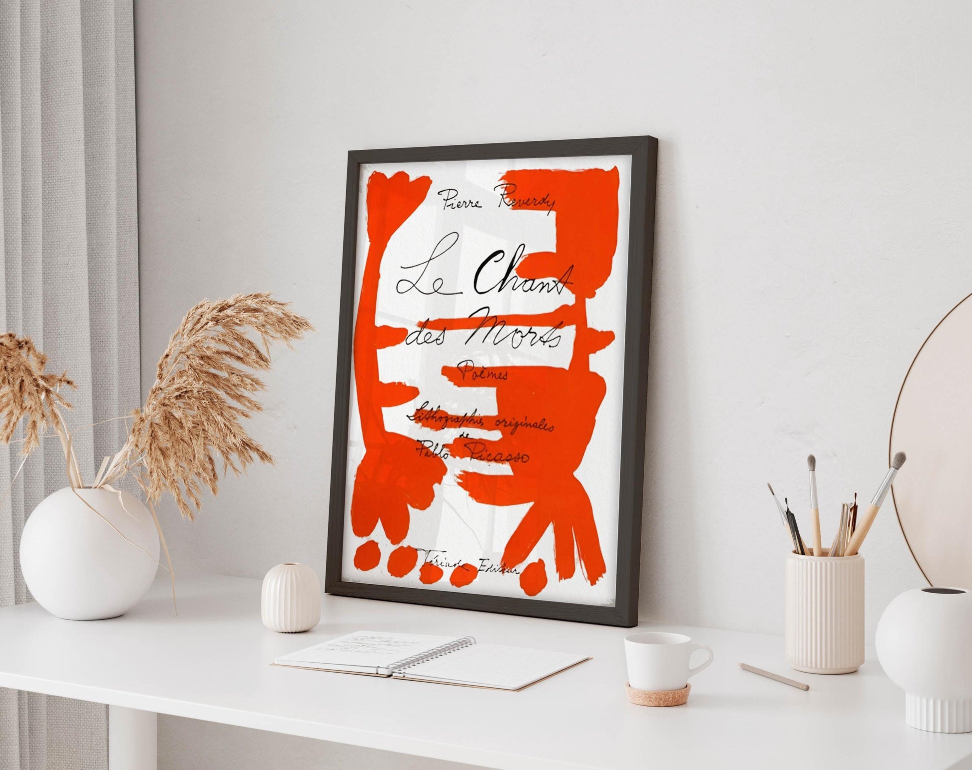 a framed poster with a red and white abstract design, placed on a white desk next to a white vase with dried flowers and a white mug.