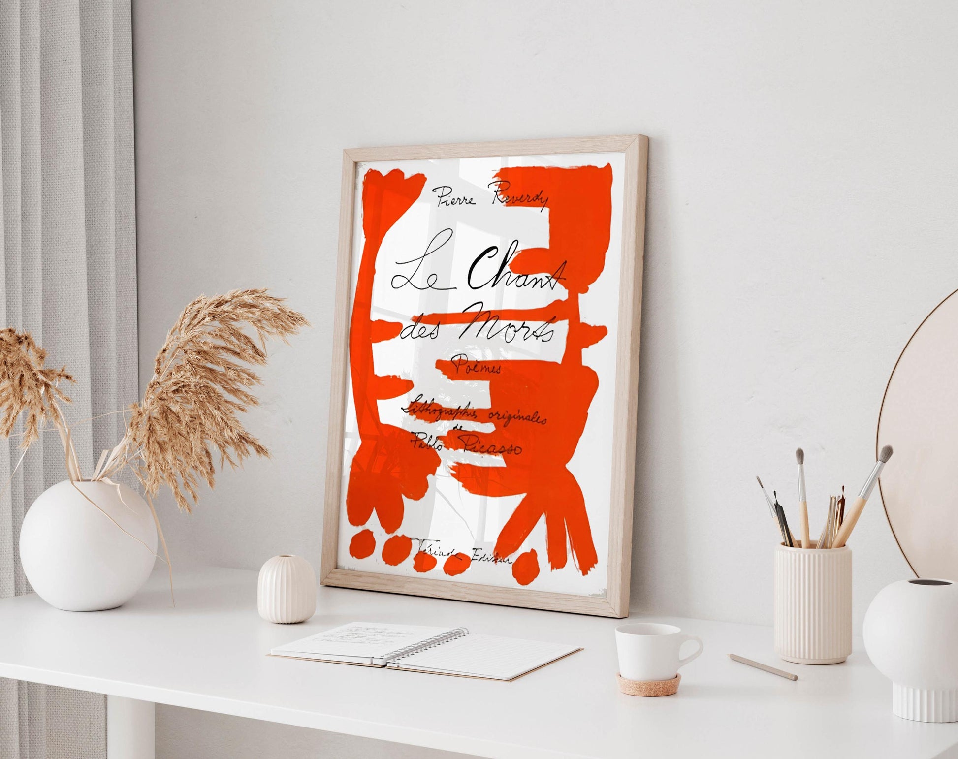 a white desk with a framed poster of an abstract red and white design hanging on the wall above it. The desk also has a notebook, a cup, and some decorative items such as a vase and a potted plant.