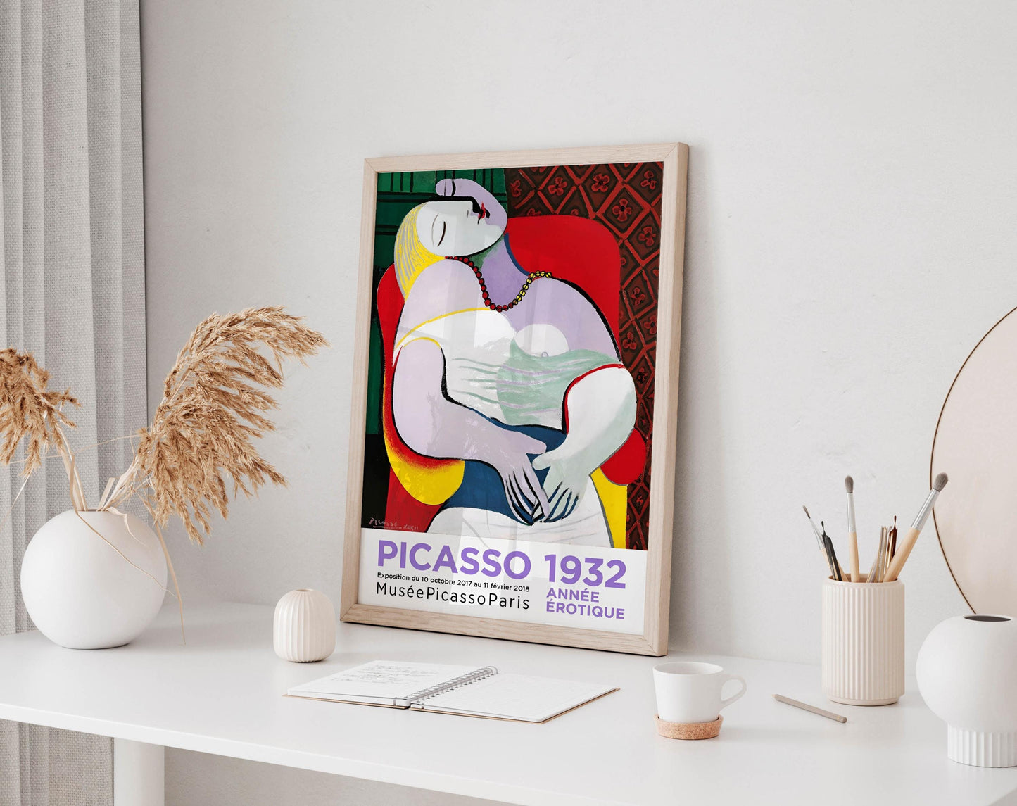 a framed poster of Picasso's cubist artwork, which is displayed on a white desk. The poster is surrounded by various objects, including a white vase, a small white candle, a white mug, and a white notebook.