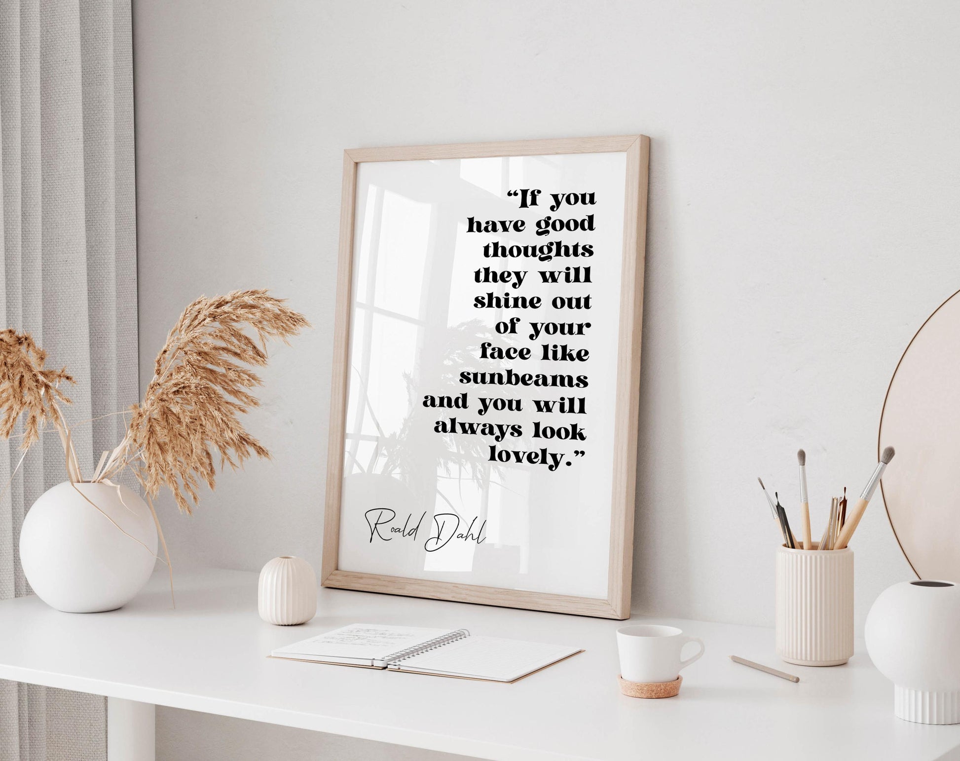 a framed quote by Ralph Dhl on a white wall, accompanied by a white desk with various items such as a notebook, a cup, and a vase with dried flowers.