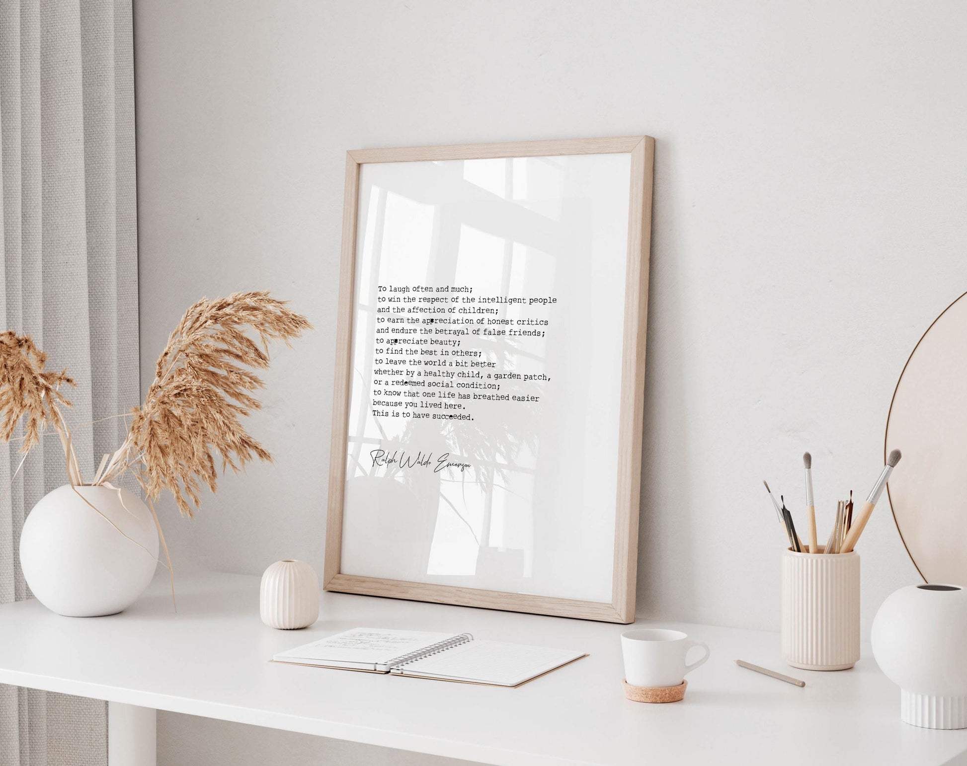 a white desk with a framed quote, a notebook, a cup, and a vase with dried flowers. The desk is set against a white wall, and there is a window in the background.