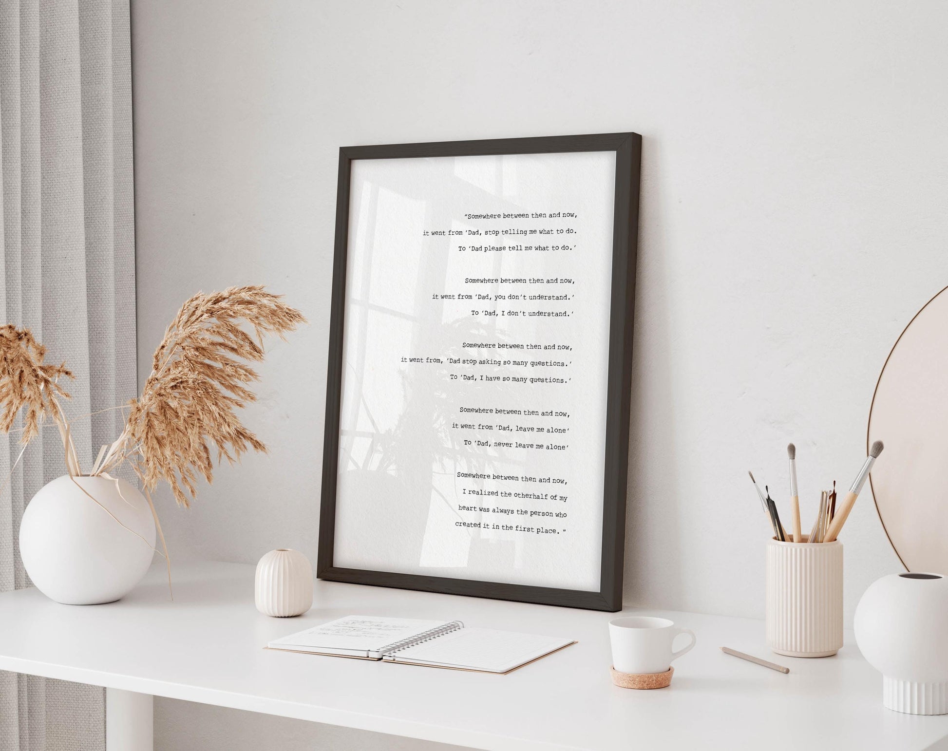 a framed poem on a white desk, surrounded by various objects such as a notebook, a cup, and a vase with dried flowers.
