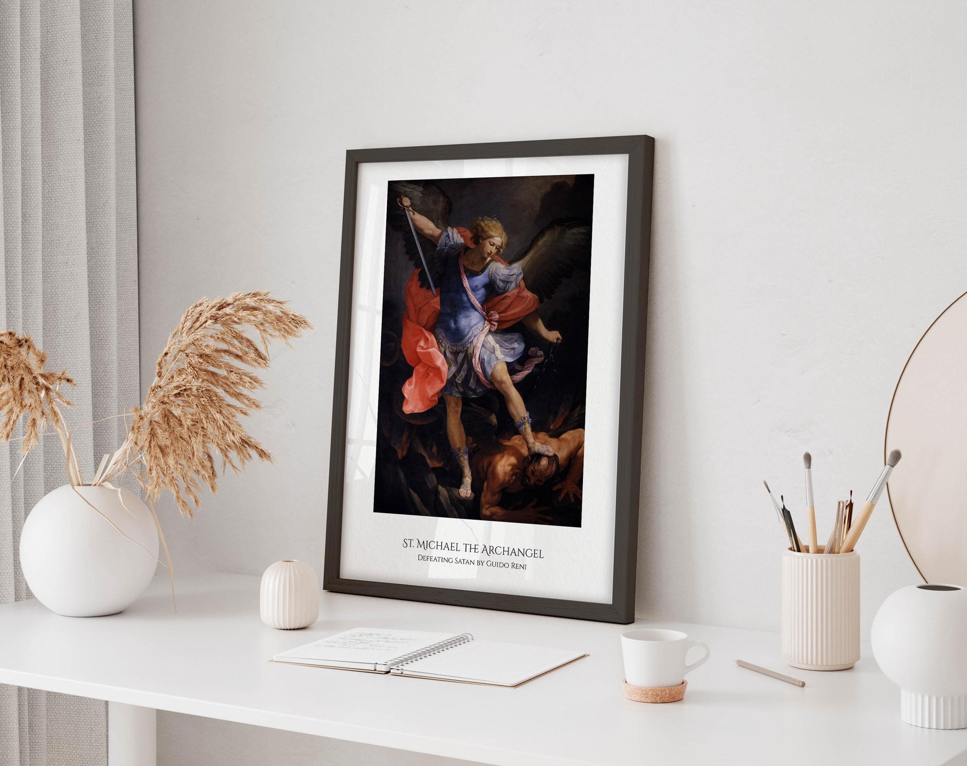 a framed painting of St. Michael the Archangel, a religious figure, standing in a dramatic pose with a sword in his hand. The painting is displayed on a white wall above a desk with various items on it, including a notebook, a cup, and a vase.