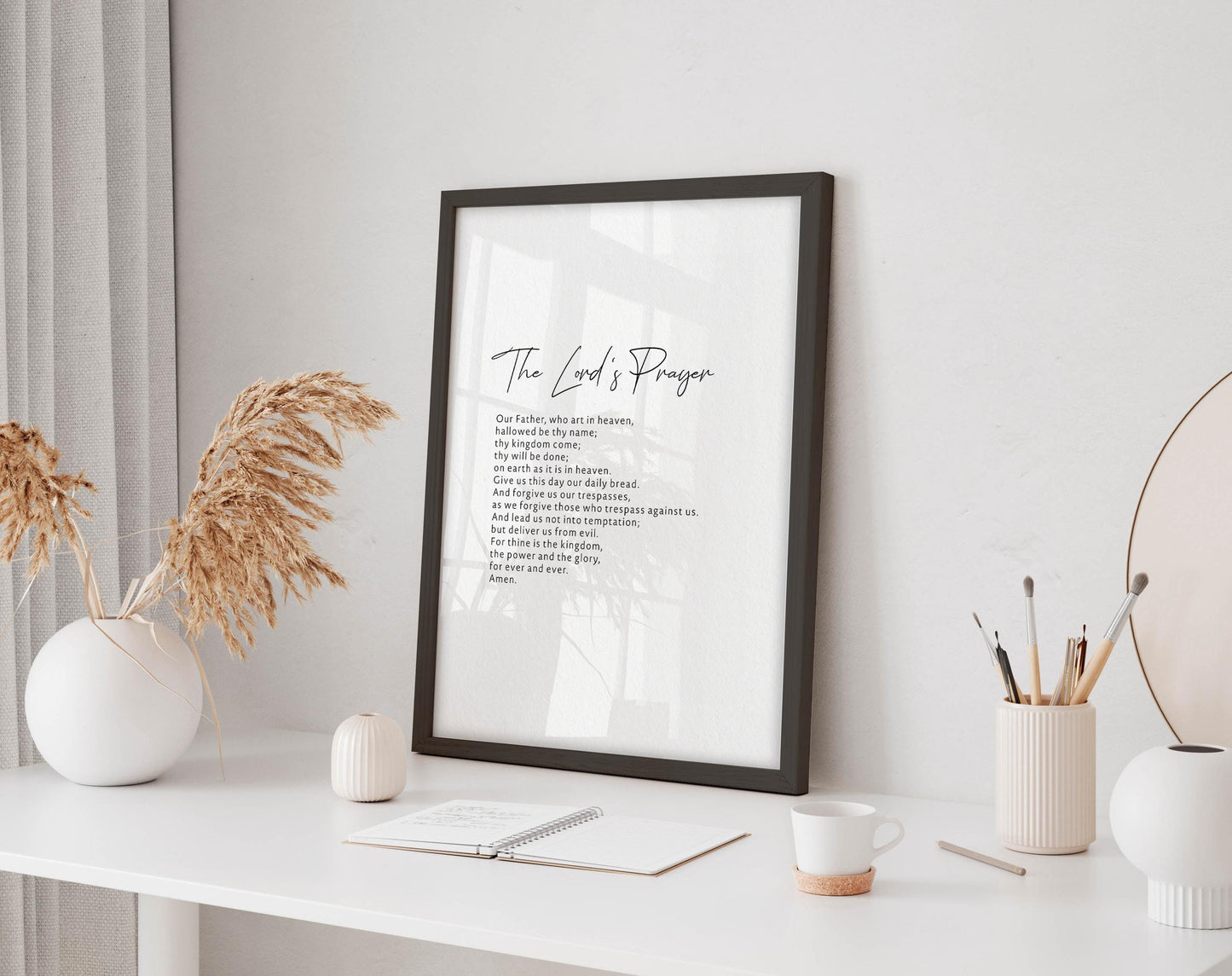 a framed prayer text on a white wall, accompanied by a white desk with various items such as a notebook, a cup, and a vase with dried flowers.
