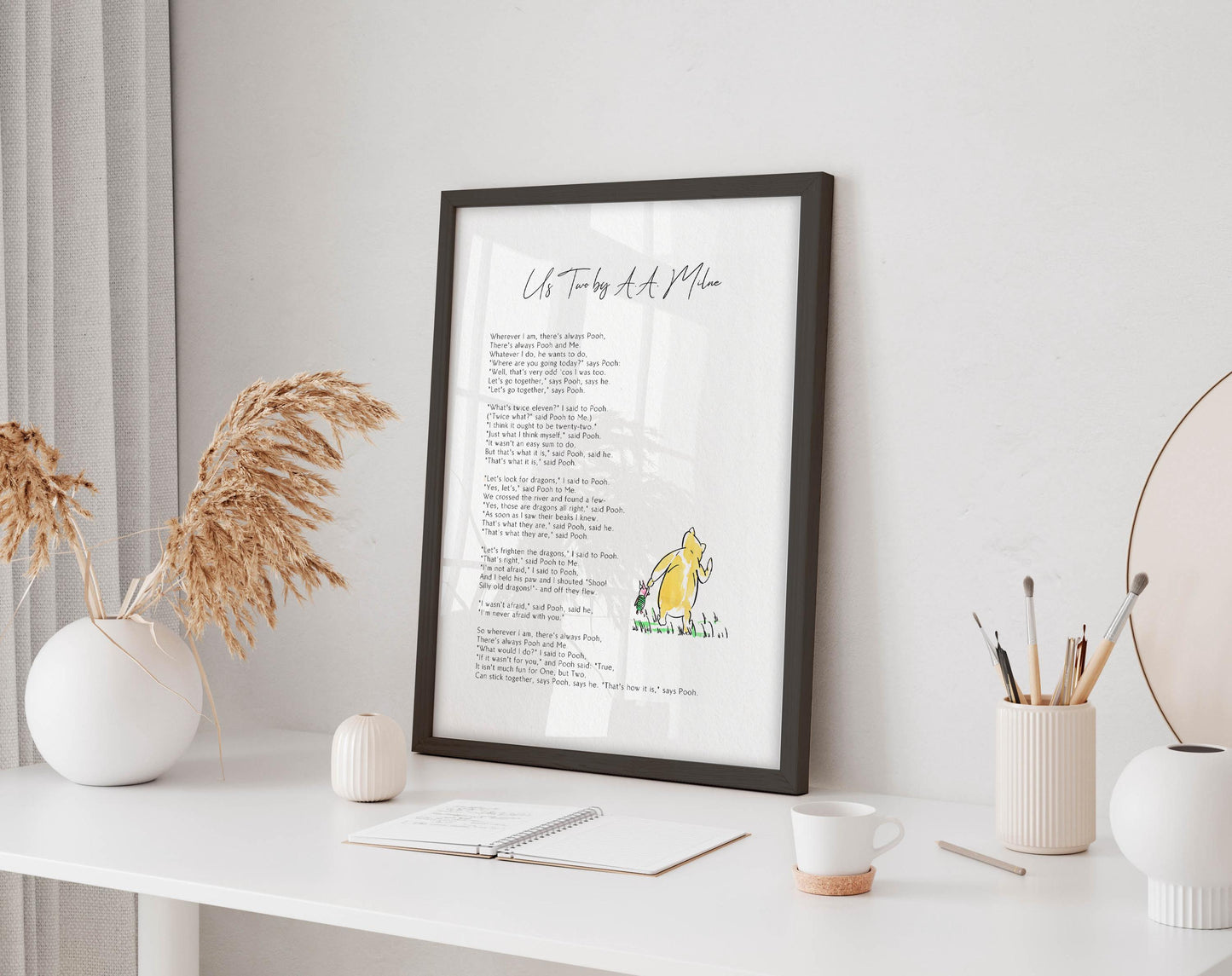 a framed poem titled "Us Too By A Moo" displayed on a white desk, accompanied by a notebook, a cup, and a vase with dried flowers.