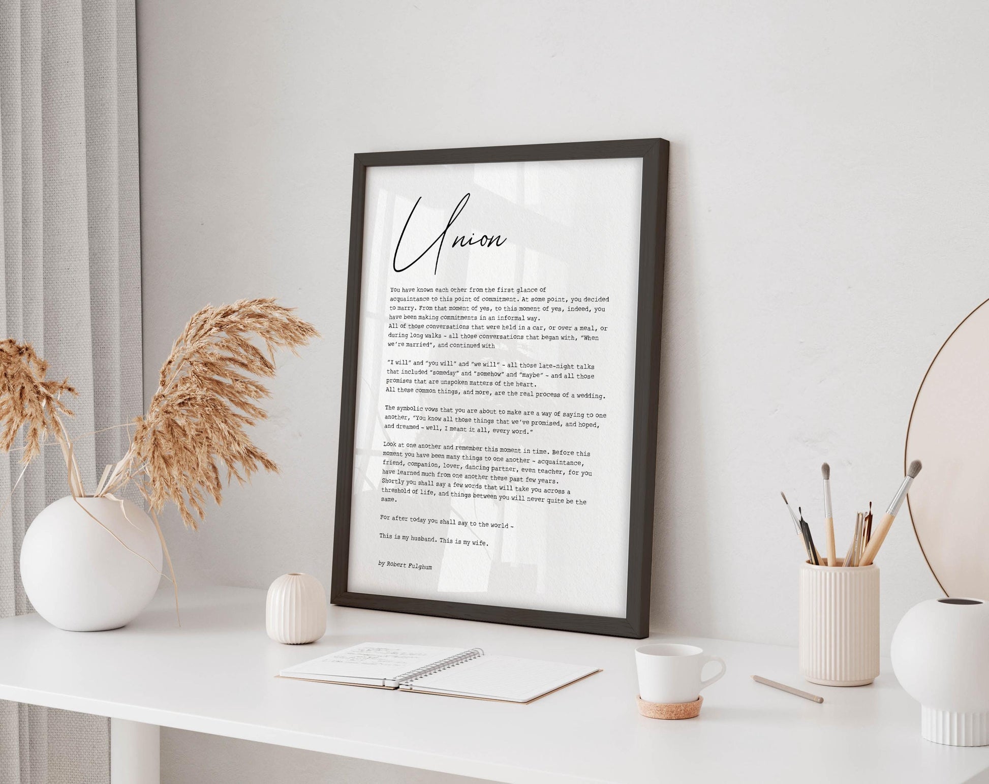a framed poster with a poem titled "Unison" displayed on a white desk, accompanied by a notebook, a cup, and a vase with dried flowers.