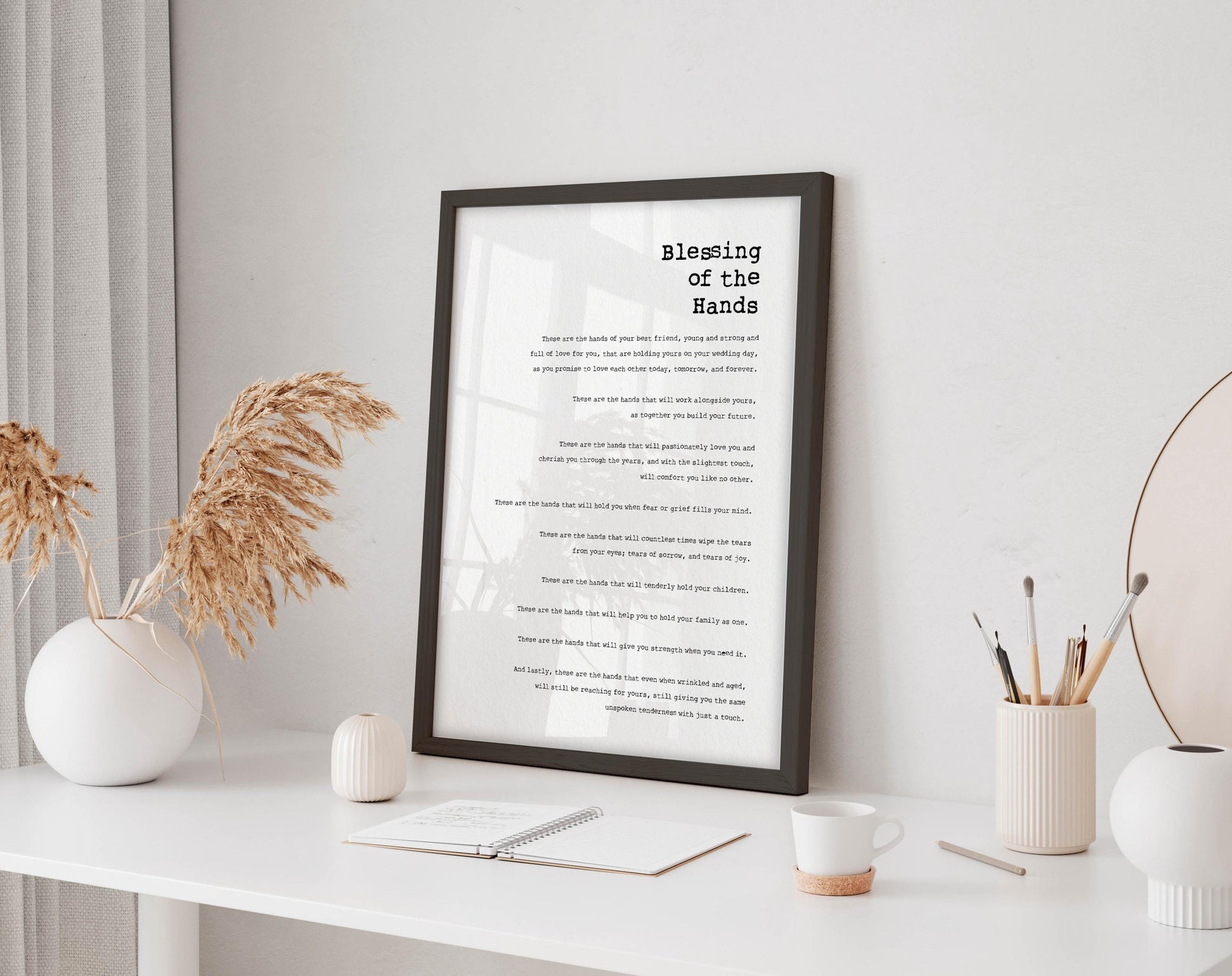 a framed poem titled "Blessing of the Hands" displayed on a white desk, with a notebook, a cup, and a vase of dried flowers nearby.