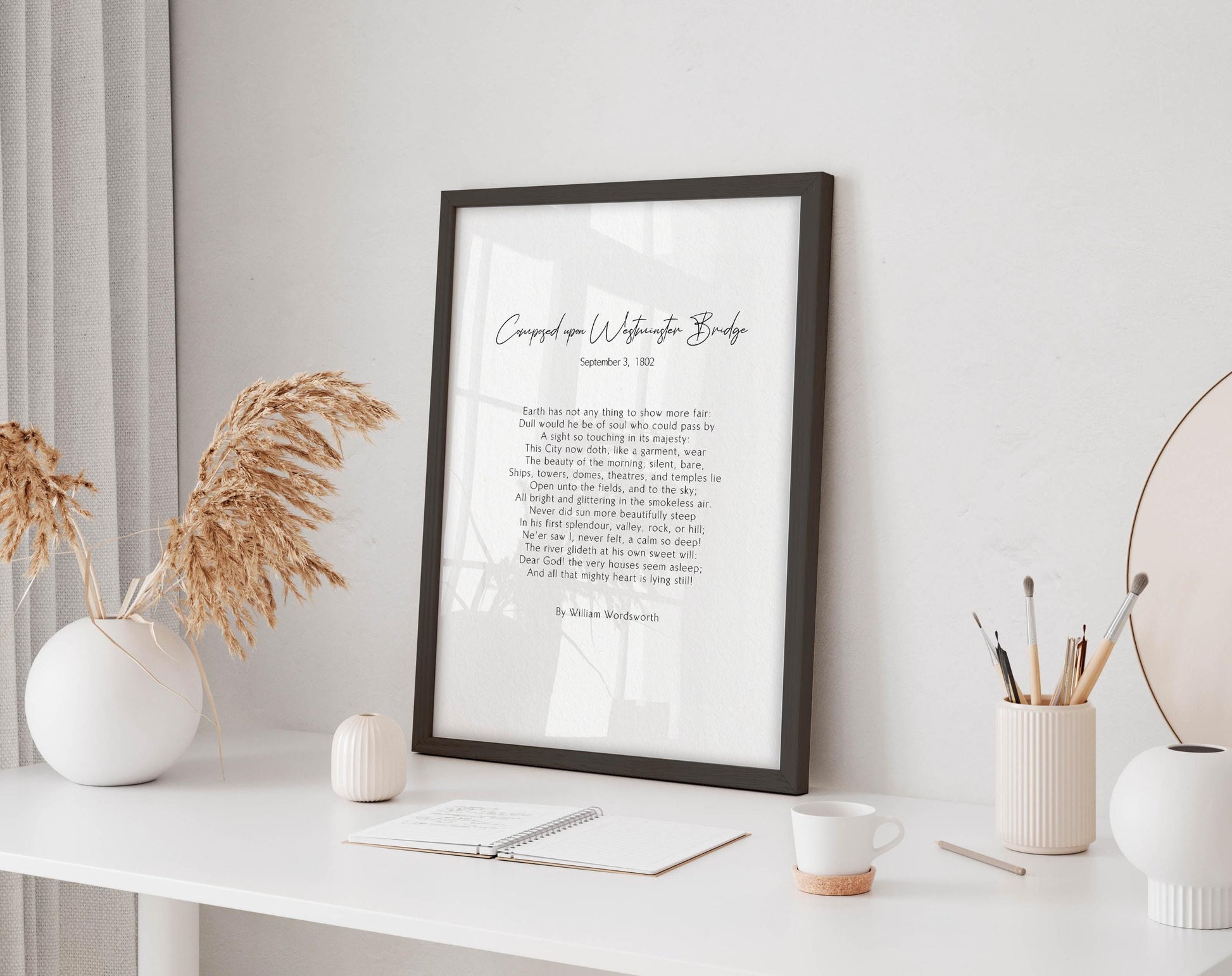 a framed poem titled "Corned on Watscot Bridge" by William Wordsworth, displayed on a white desk with a notebook, pen, and a cup of coffee. The desk is set against a white wall, with a vase of dried flowers and a mirror adding to the decor.