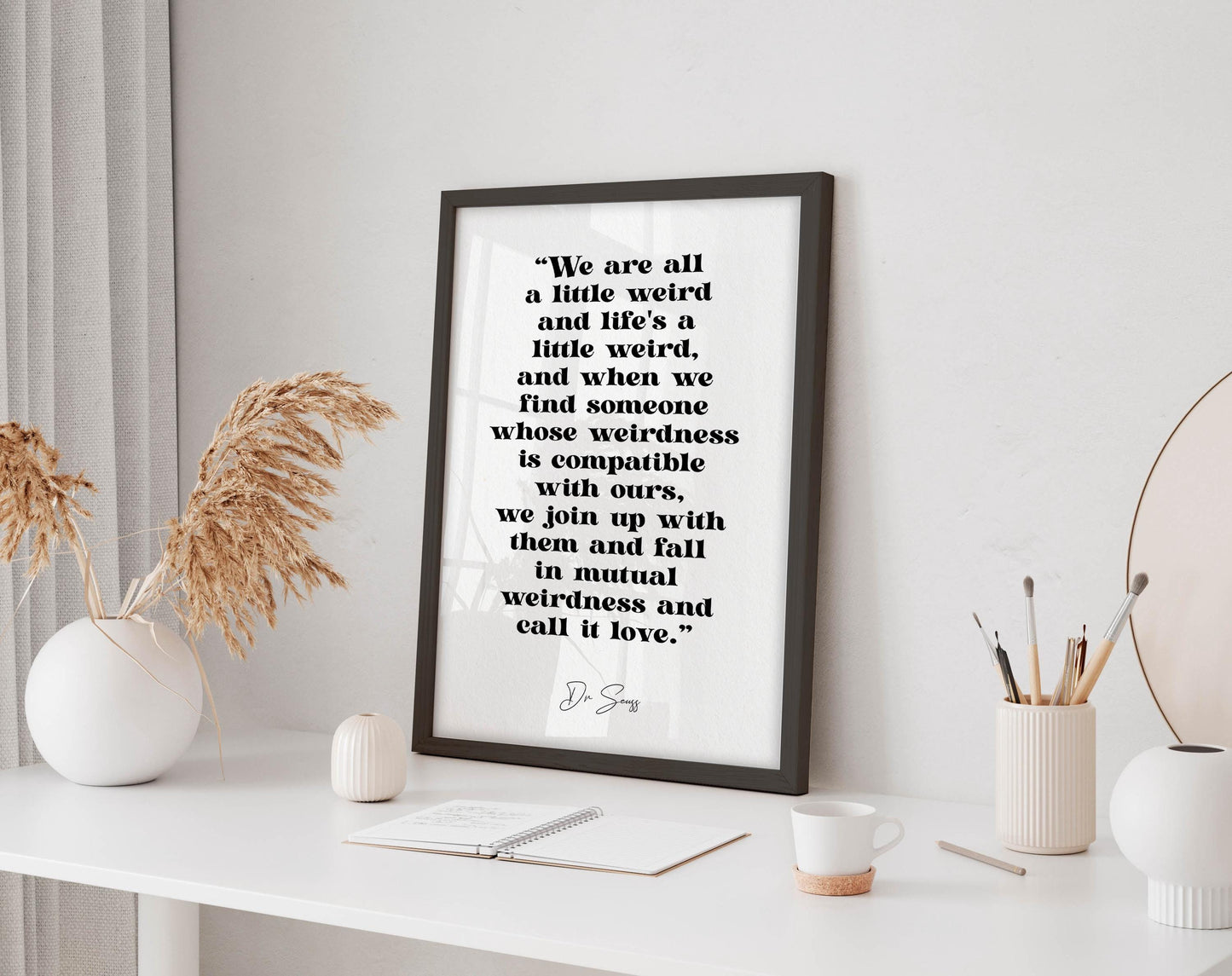 a framed quote by D.S. Jain, "We are all a little weird and life's a little weird, and when we find someone whose weirdness is compatible with ours, we join up with them and fall in mutual weirdness and call it love." The quote is displayed on a white wall, accompanied by a small white vase with dried flowers, a white mug, and a small white notebook.