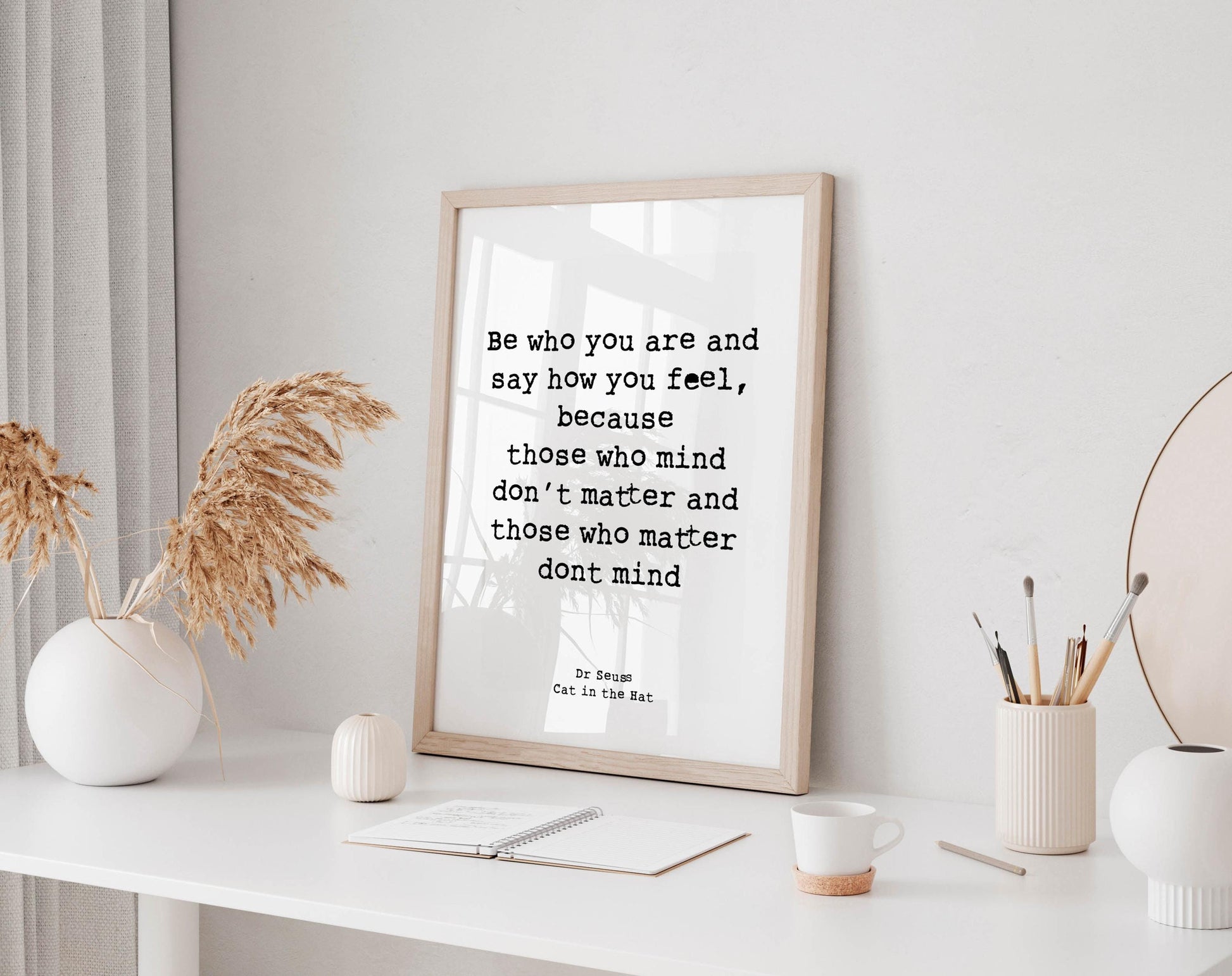 a framed quote on a white wall, surrounded by various objects such as a notebook, a cup, and a vase with dried flowers.