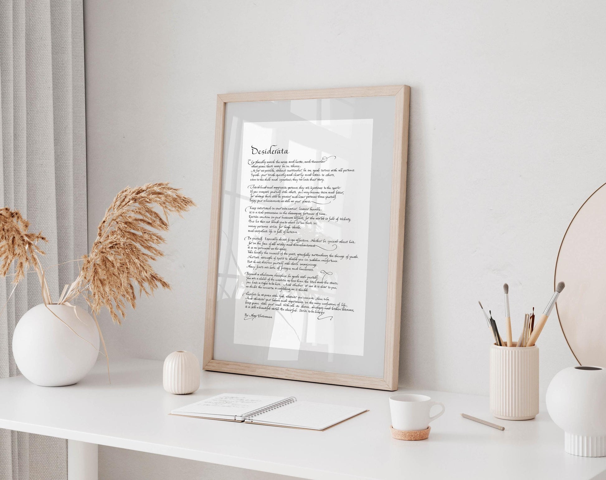 a framed text on a white desk, surrounded by various objects such as a cup, a notebook, and a vase with dried flowers.