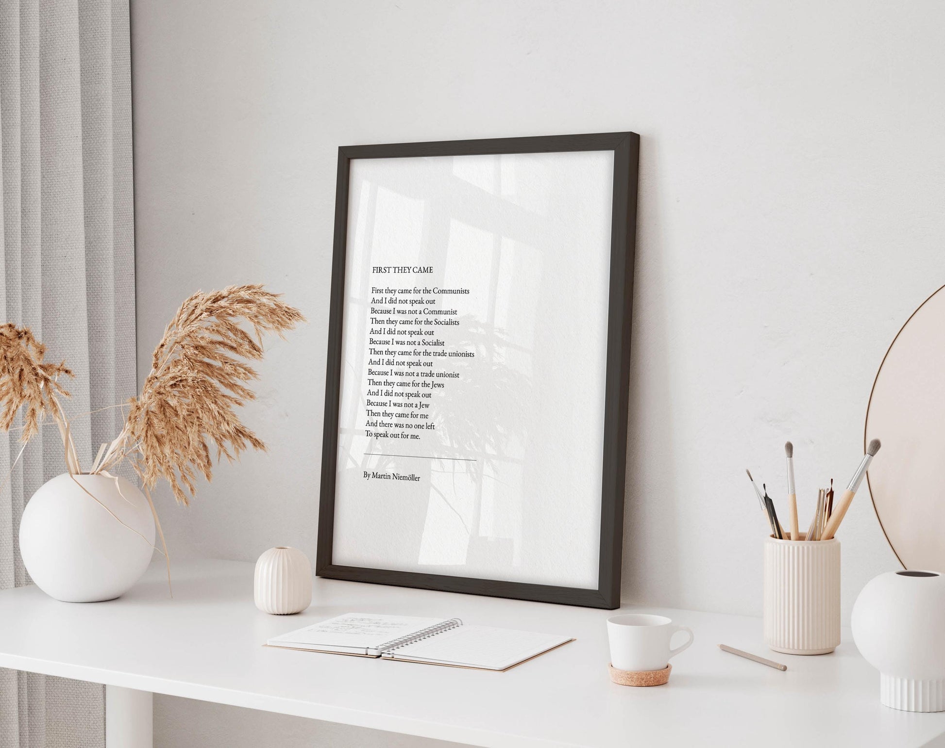 a framed poem titled "First They Came" displayed on a white desk, accompanied by a notebook, a cup, and a vase with dried flowers.