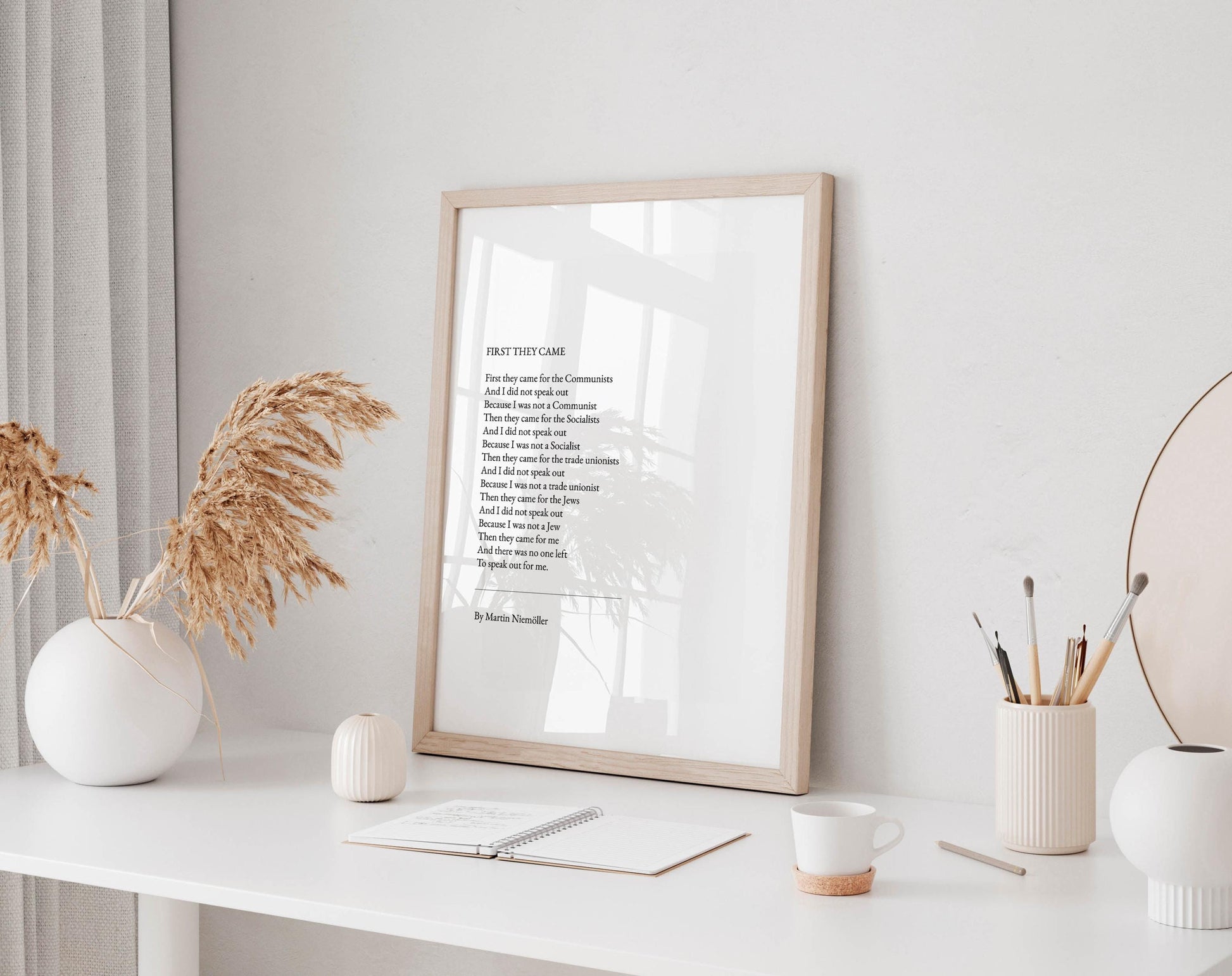 a framed poem titled "First They Came" displayed on a white desk, accompanied by a notebook, a cup, and a vase with dried flowers.
