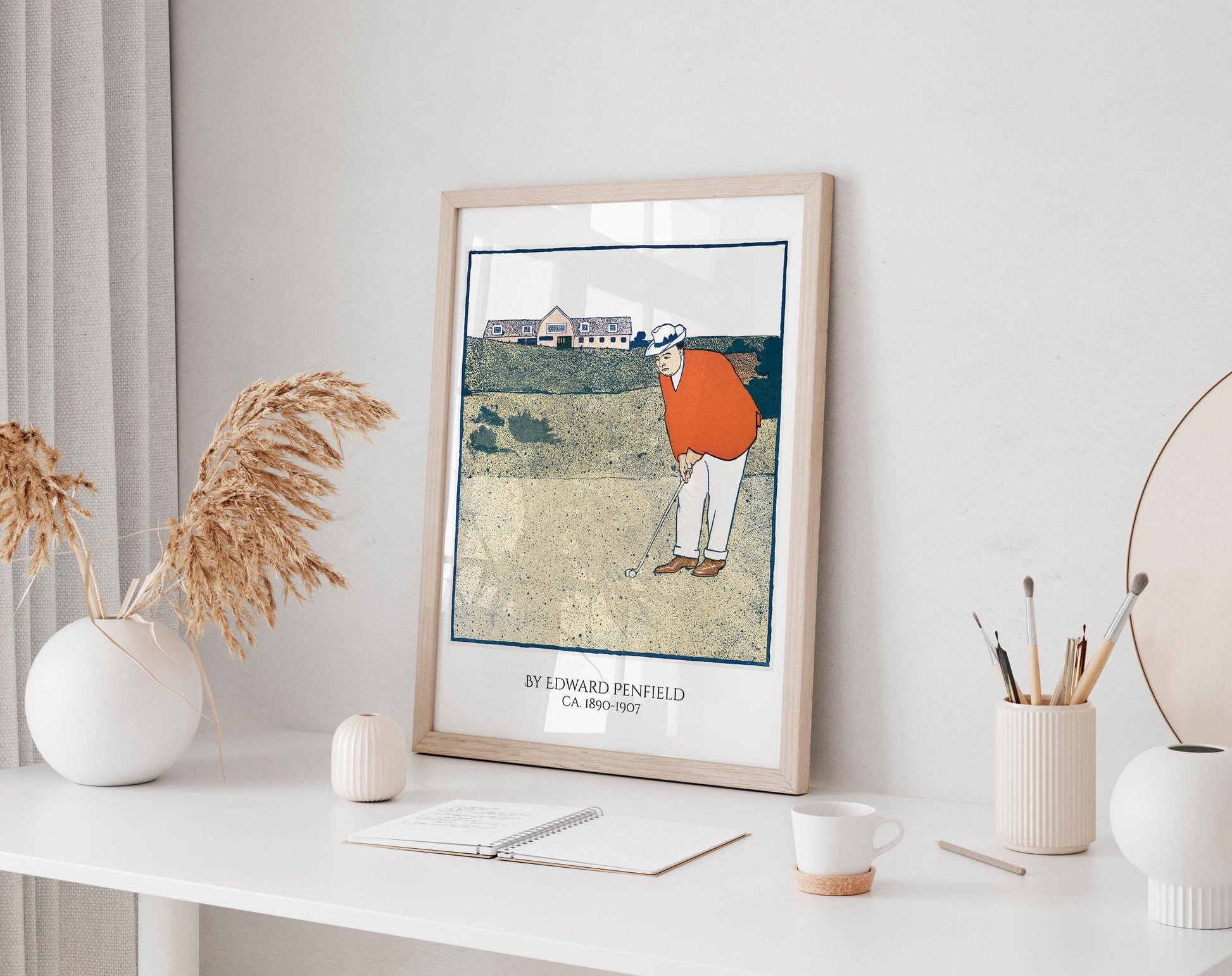 a framed illustration of a golfer on a golf course, with a vase of dried flowers and a cup of coffee on a desk in the foreground.