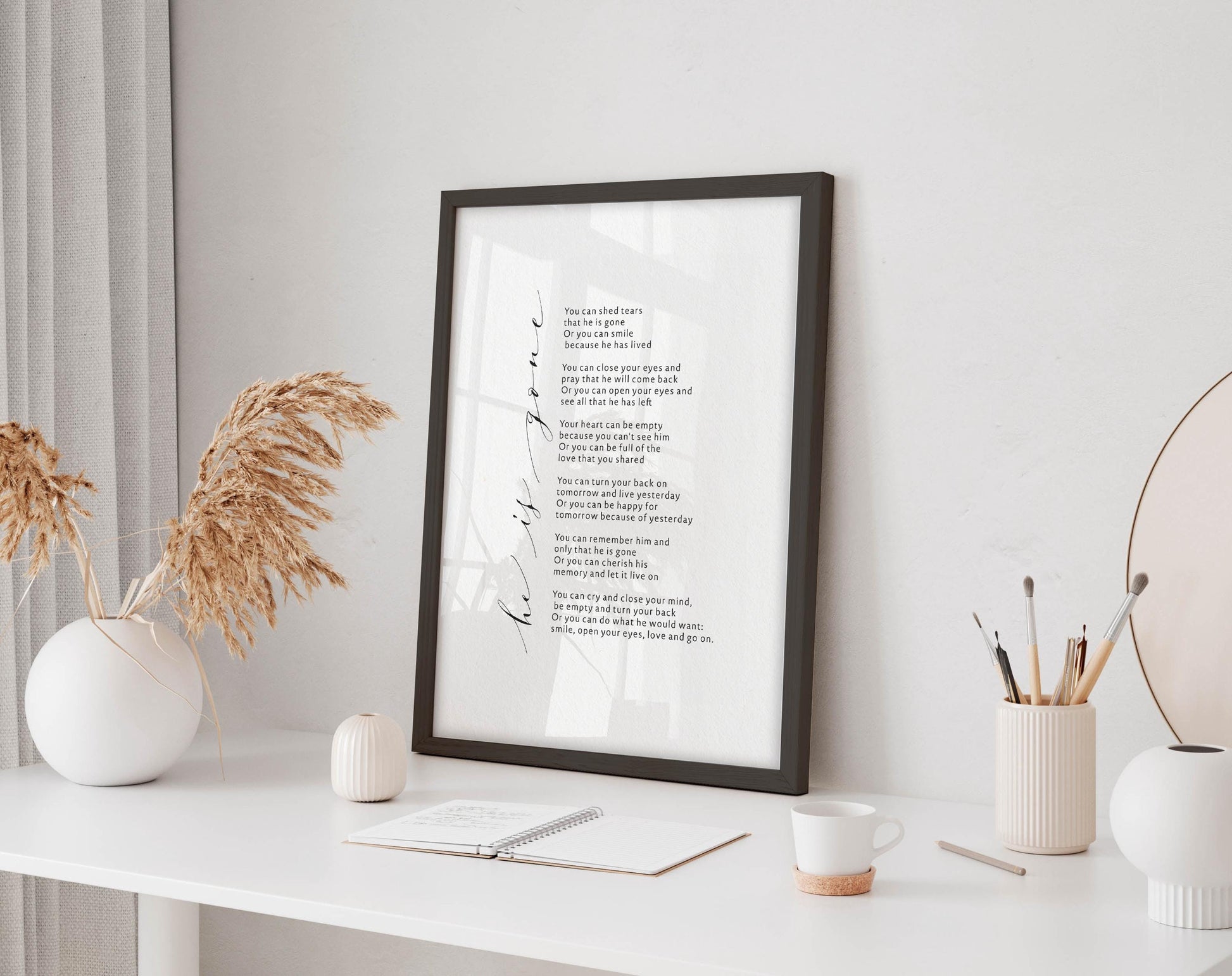 a framed poem on a white desk, surrounded by various objects such as a notebook, a cup, and a vase with dried flowers.