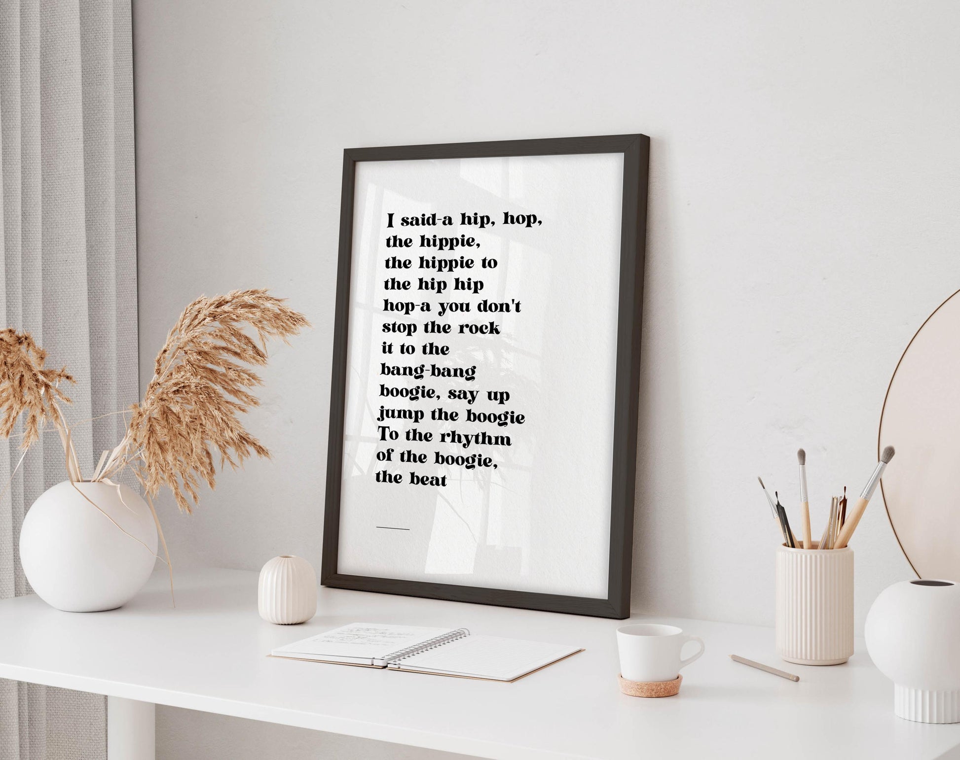 a framed quote on a white wall, accompanied by a white desk with various items such as a coffee cup, a notebook, and a vase with dried flowers.