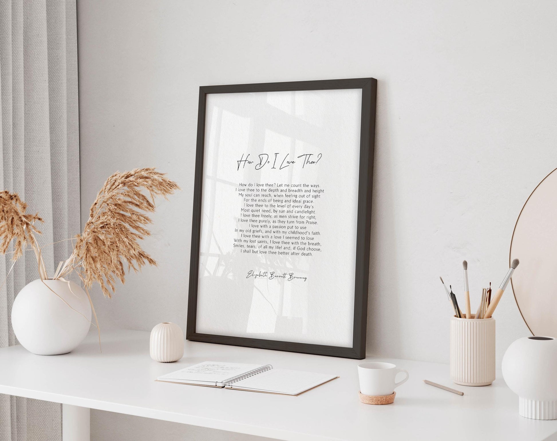 a framed quote on a white desk, surrounded by various objects such as a white vase, a notebook, a cup, and a pen holder.