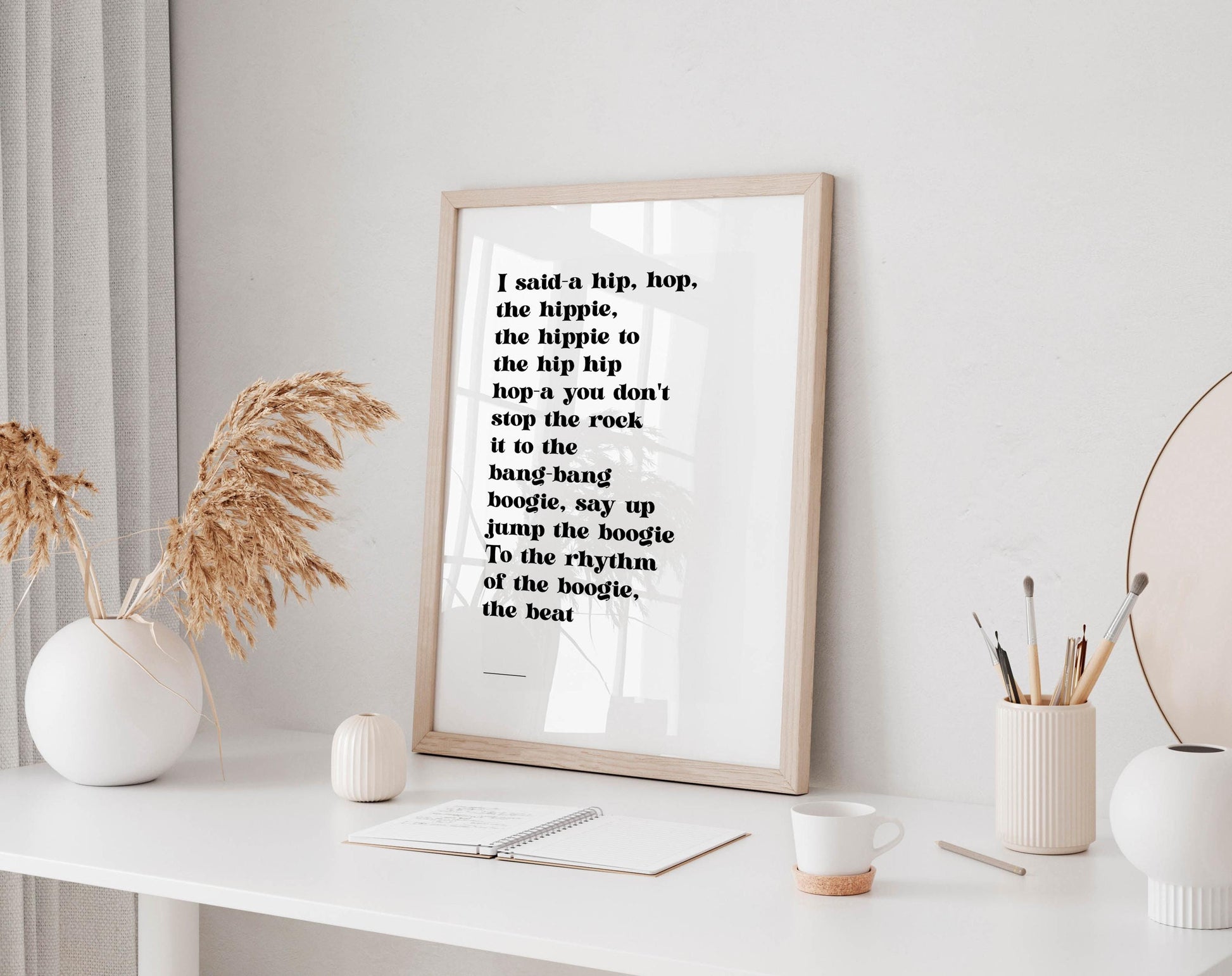 a framed quote on a white wall, accompanied by a white desk with various items such as a coffee cup, a notebook, and a vase with dried flowers.