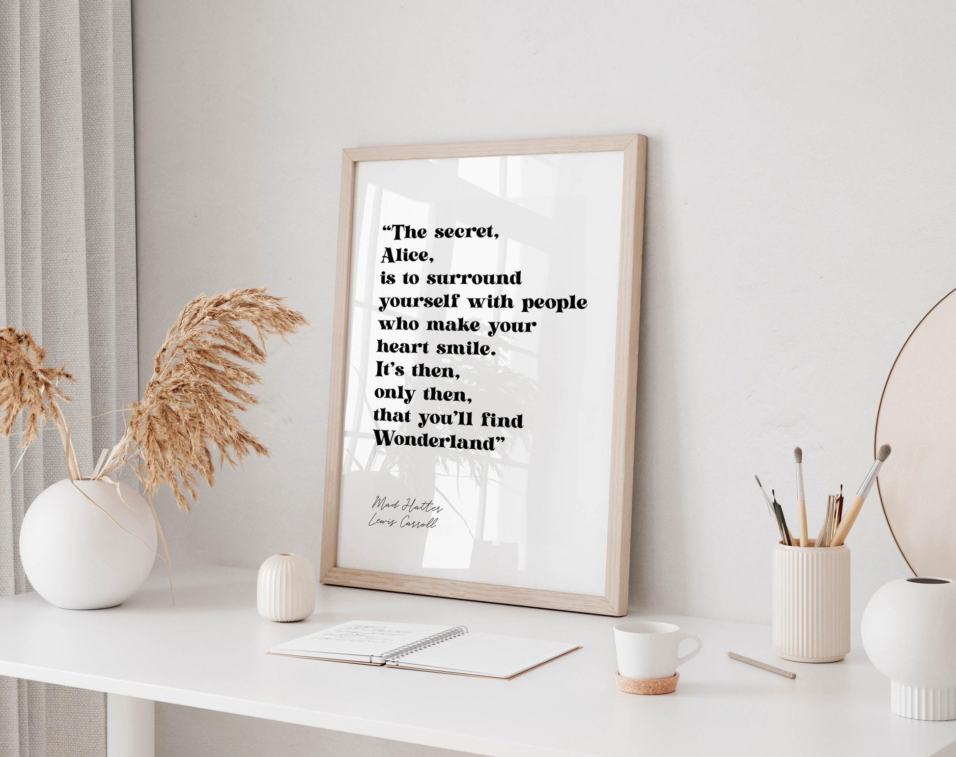a framed quote by Lewis Carroll, "The secret, Alice, is to surround yourself with people who make your heart smile. It's then, only then, that you'll find Wonderland." The quote is displayed on a white wall, with a white desk and various objects such as a vase, a cup, and a notebook in the foreground.