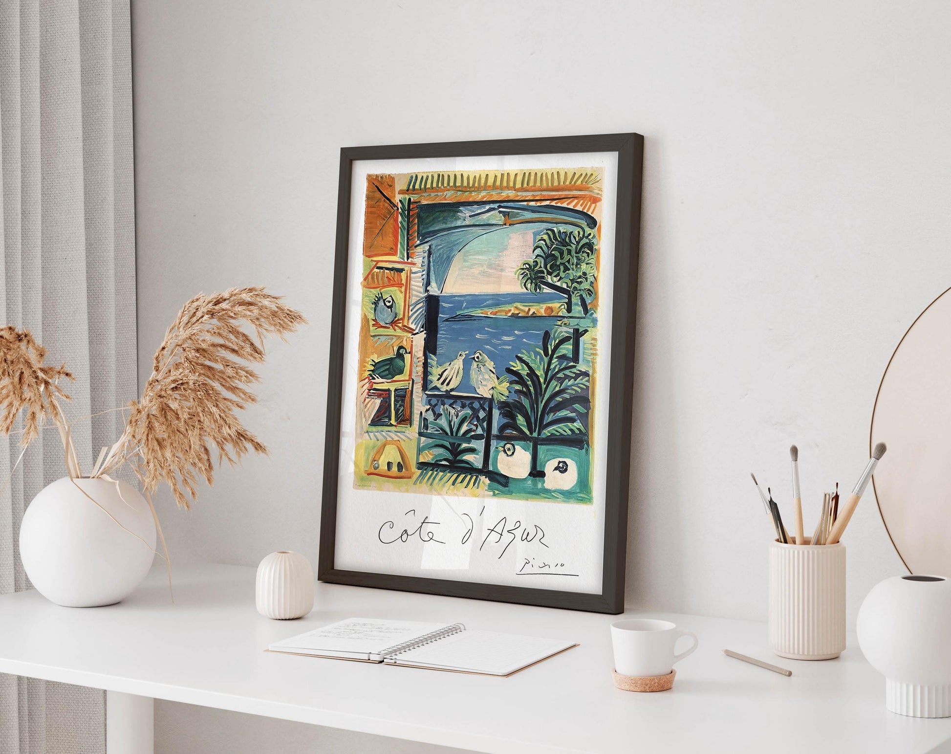 a framed painting of a tropical landscape with palm trees, birds, and a body of water, displayed on a white shelf. The painting is surrounded by various decorative items, including a vase with dried flowers, a small white candle, and a white mug.