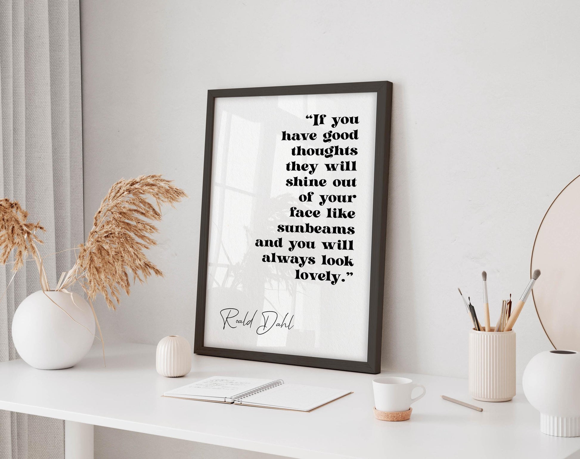 a framed quote by Ralph Dhl on a white wall, accompanied by a desk with a notebook, a cup, and a vase with dried flowers.