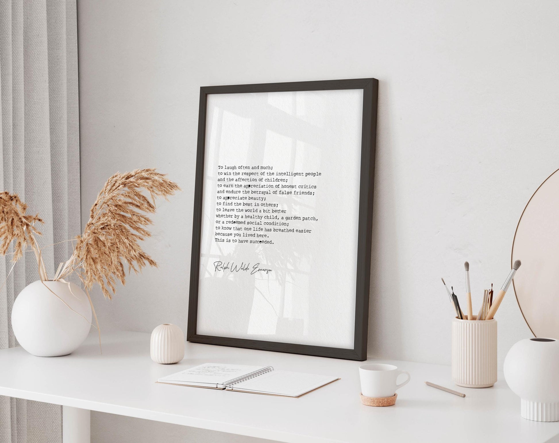 a framed quote on a white desk, surrounded by various objects such as a notebook, a cup, and a vase with dried flowers.