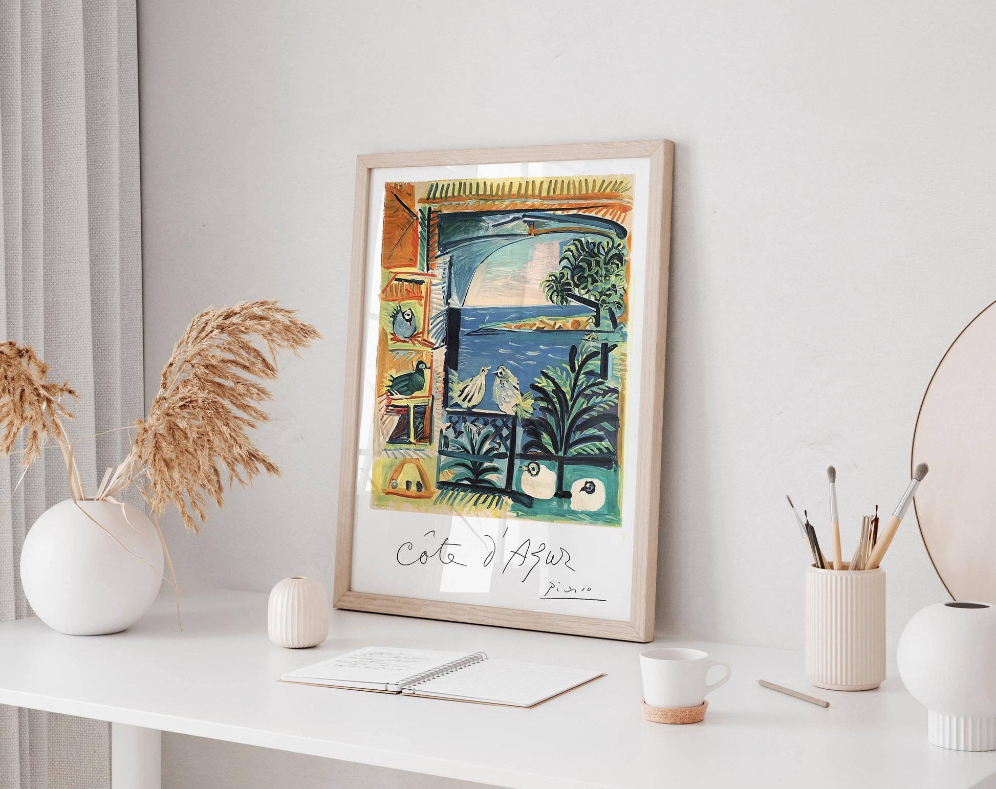 a framed painting of a tropical landscape with palm trees, birds, and a body of water, displayed on a white wall above a desk with various items on it.