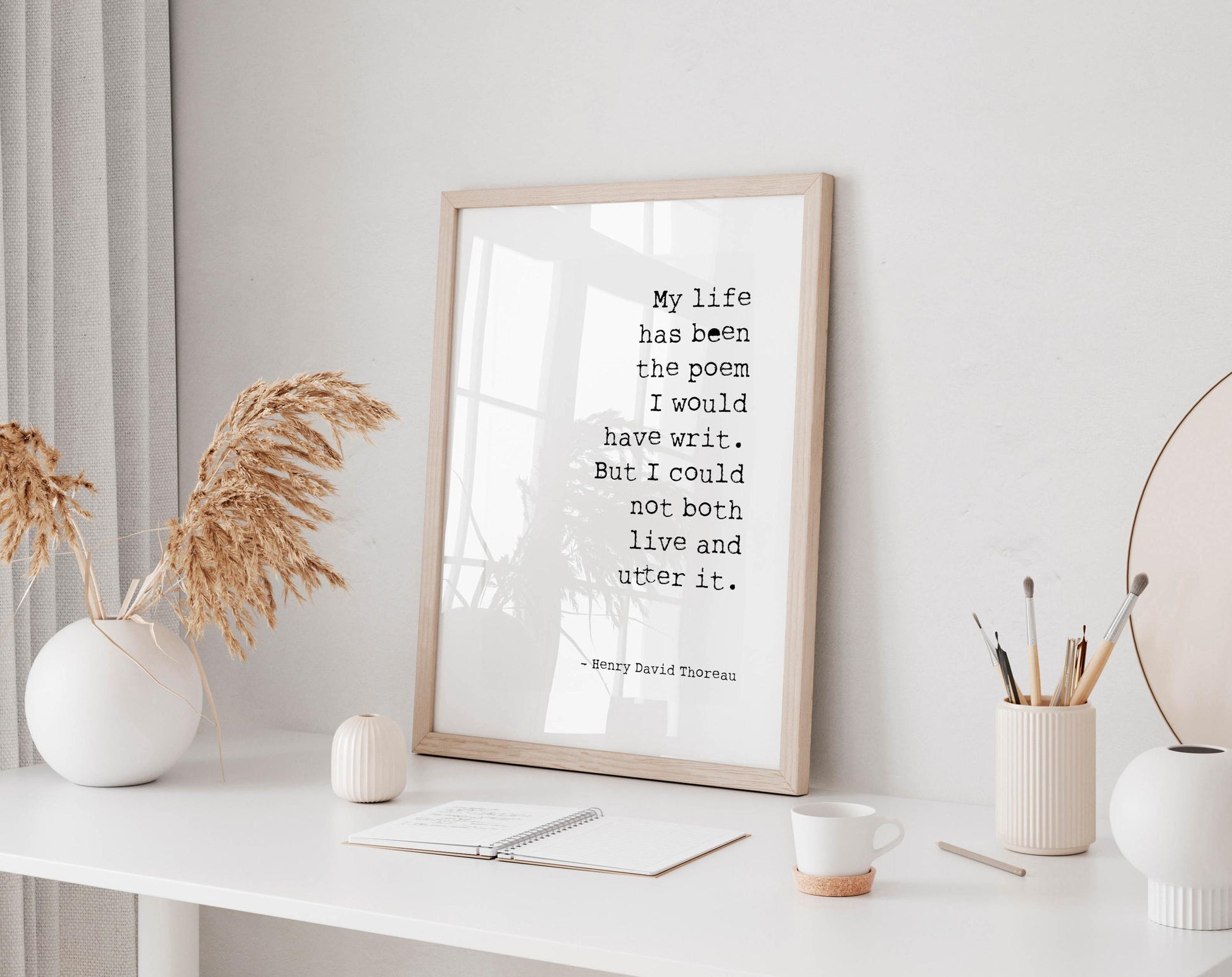 a framed quote on a white desk, surrounded by various objects such as a notebook, a cup, and a vase with dried flowers.
