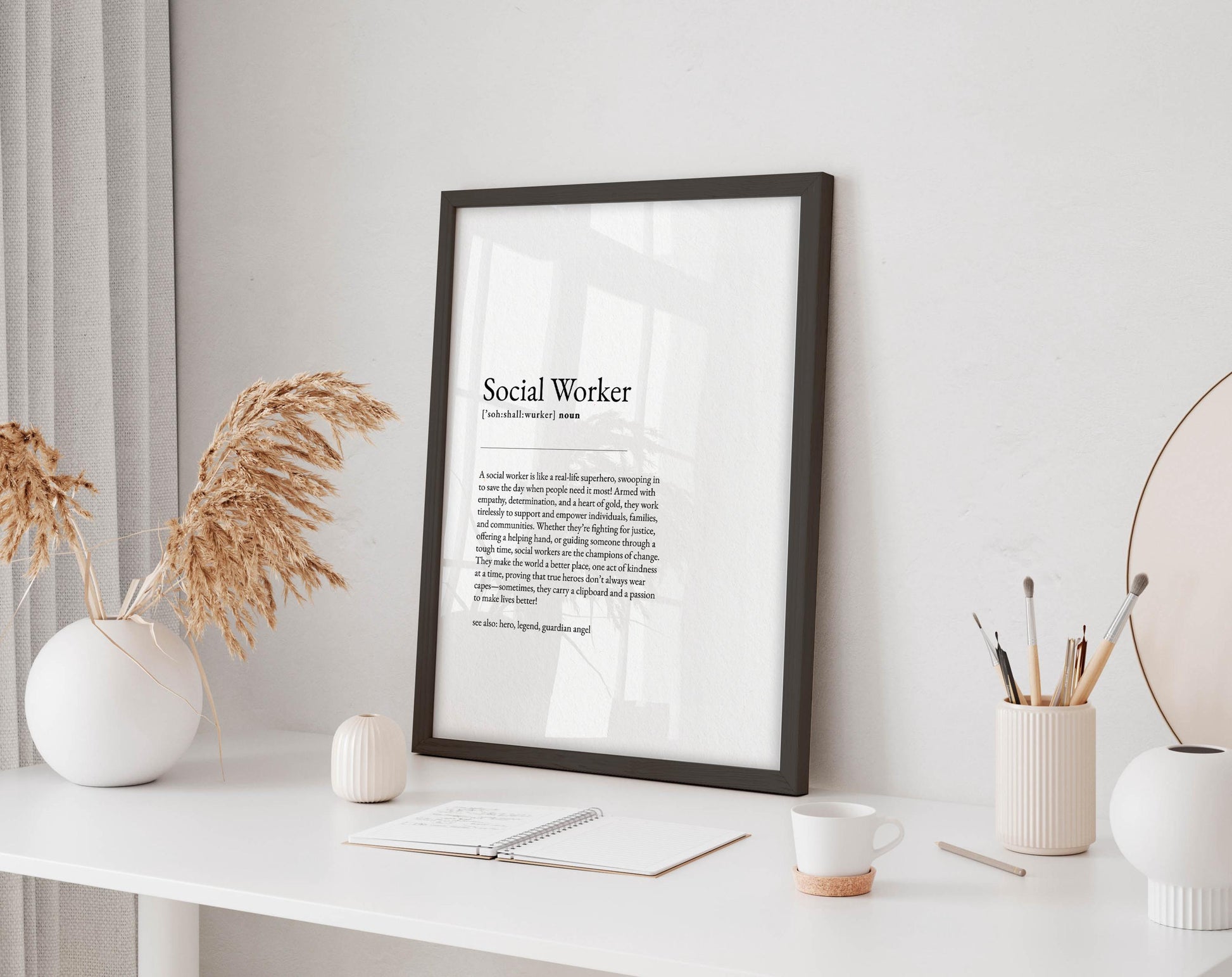 a framed definition of a social worker, a white desk with various items such as a notebook, a cup, a vase with dried flowers, and a mirror.