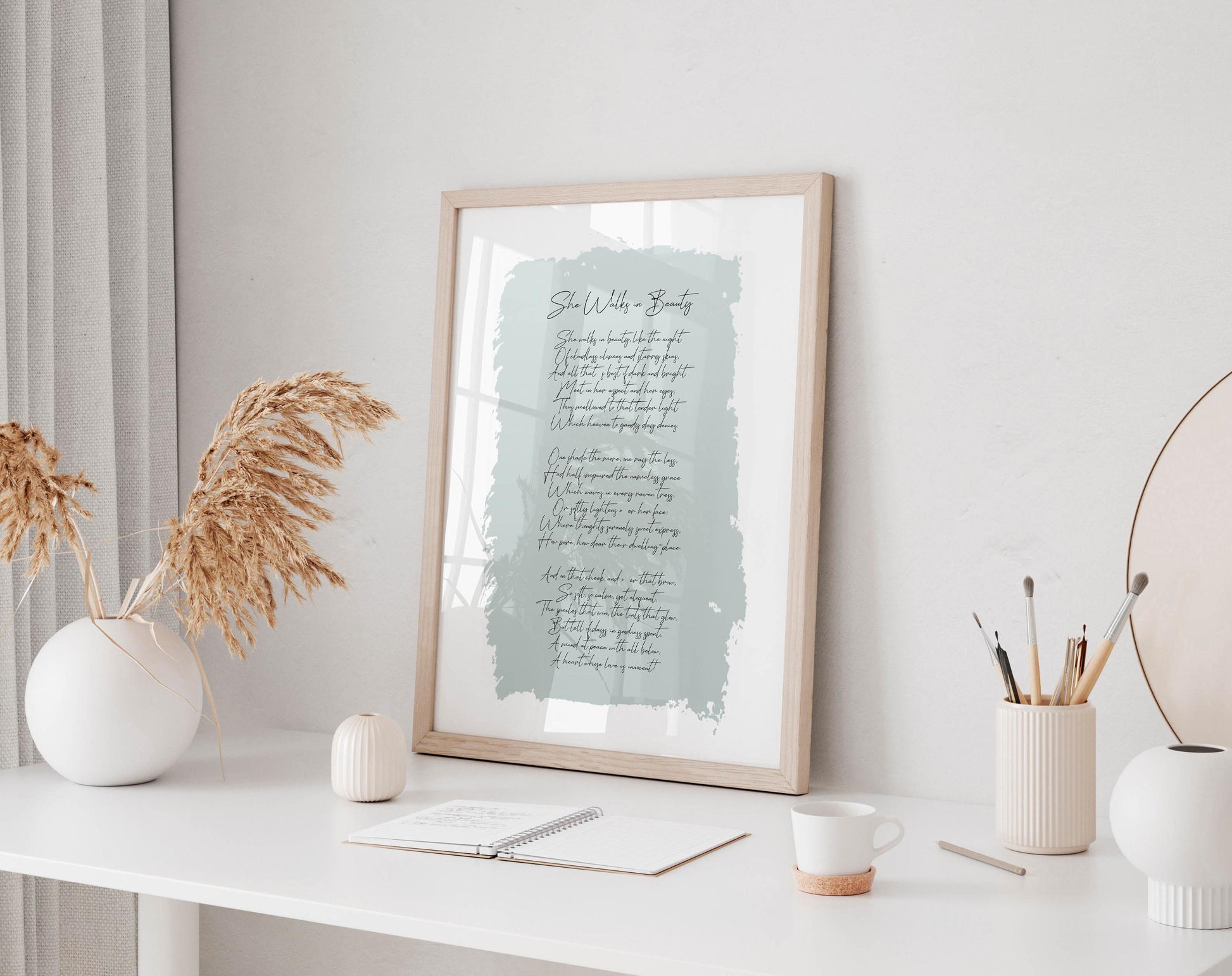 a white desk with a framed print of a poem, a white vase with dried flowers, a white mug, a white notebook, and a white candle.