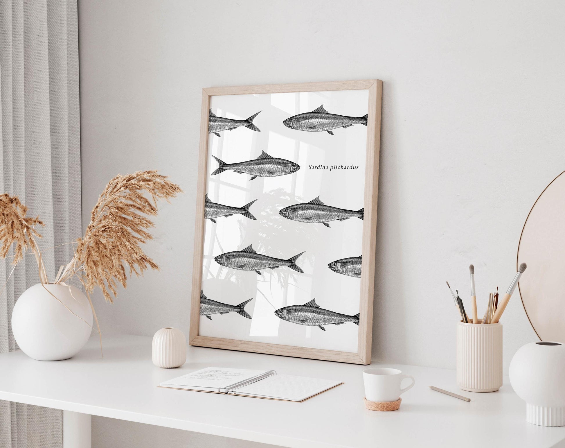 a white desk with a framed poster of fish, a notebook, a cup, and a vase with dried flowers.