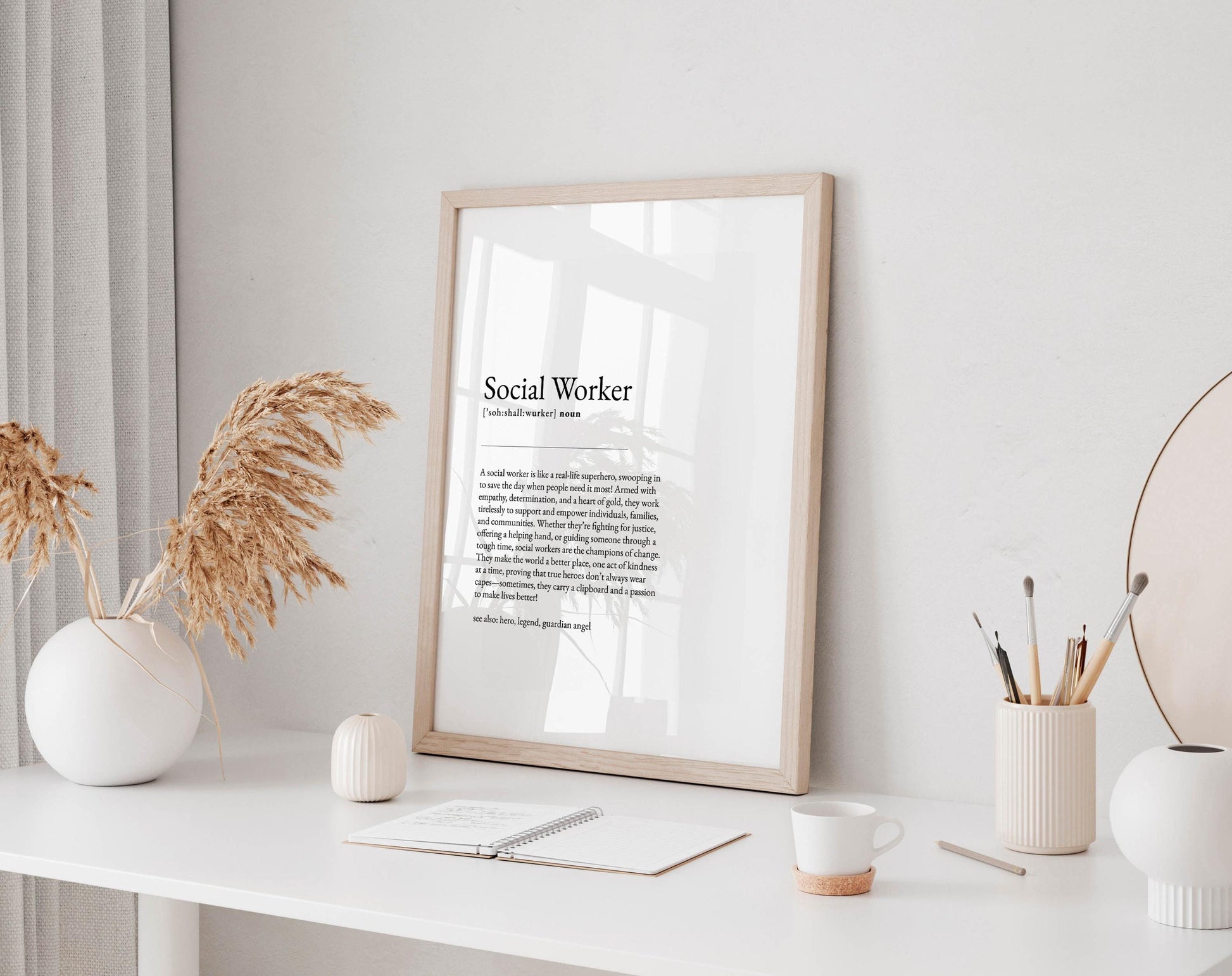 a desk with a framed definition of a social worker, a white vase with dried flowers, a white mug, a notebook, and a small white candle.