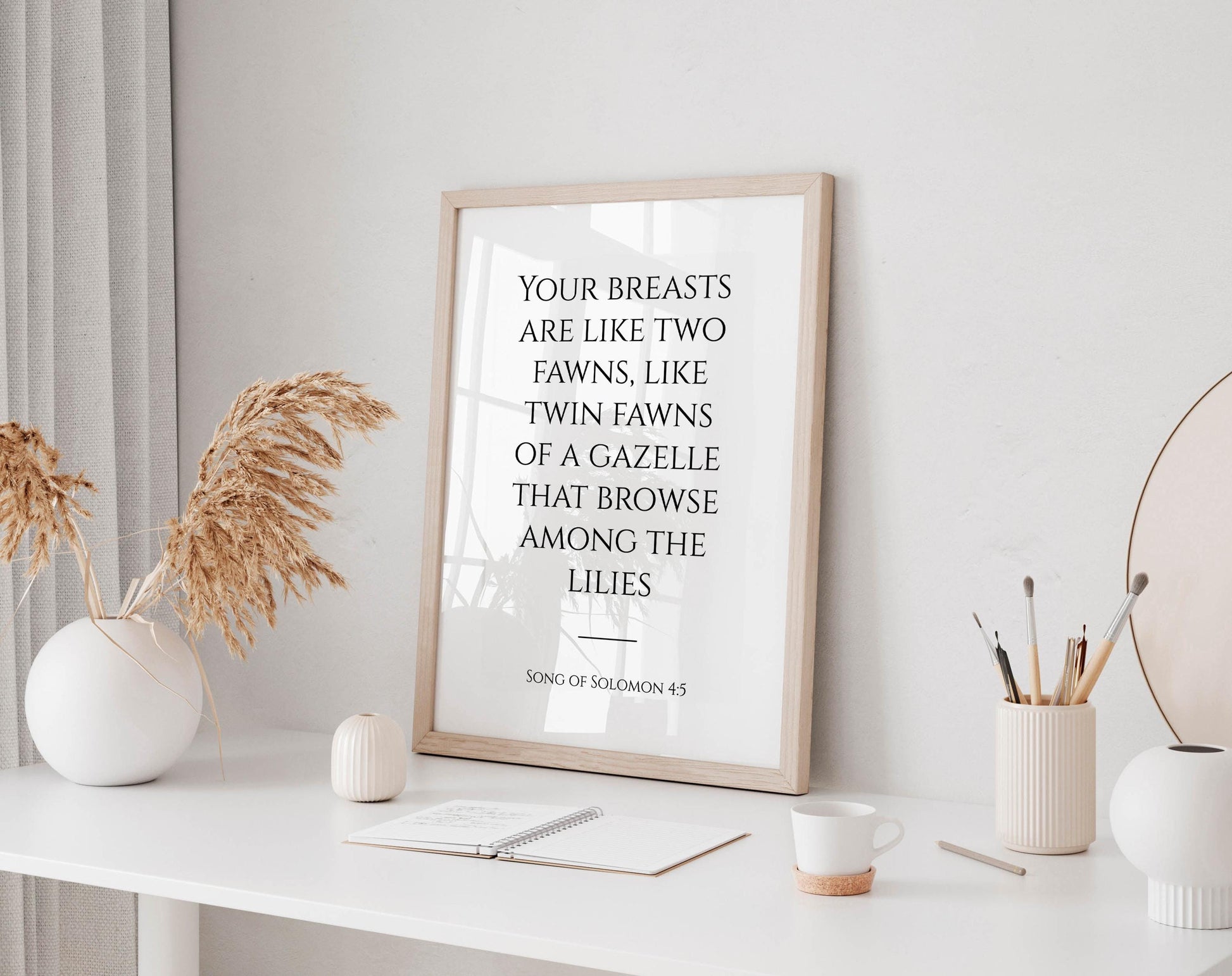 a framed quote on a white desk, surrounded by various objects such as a notebook, a cup, and a vase with dried flowers.