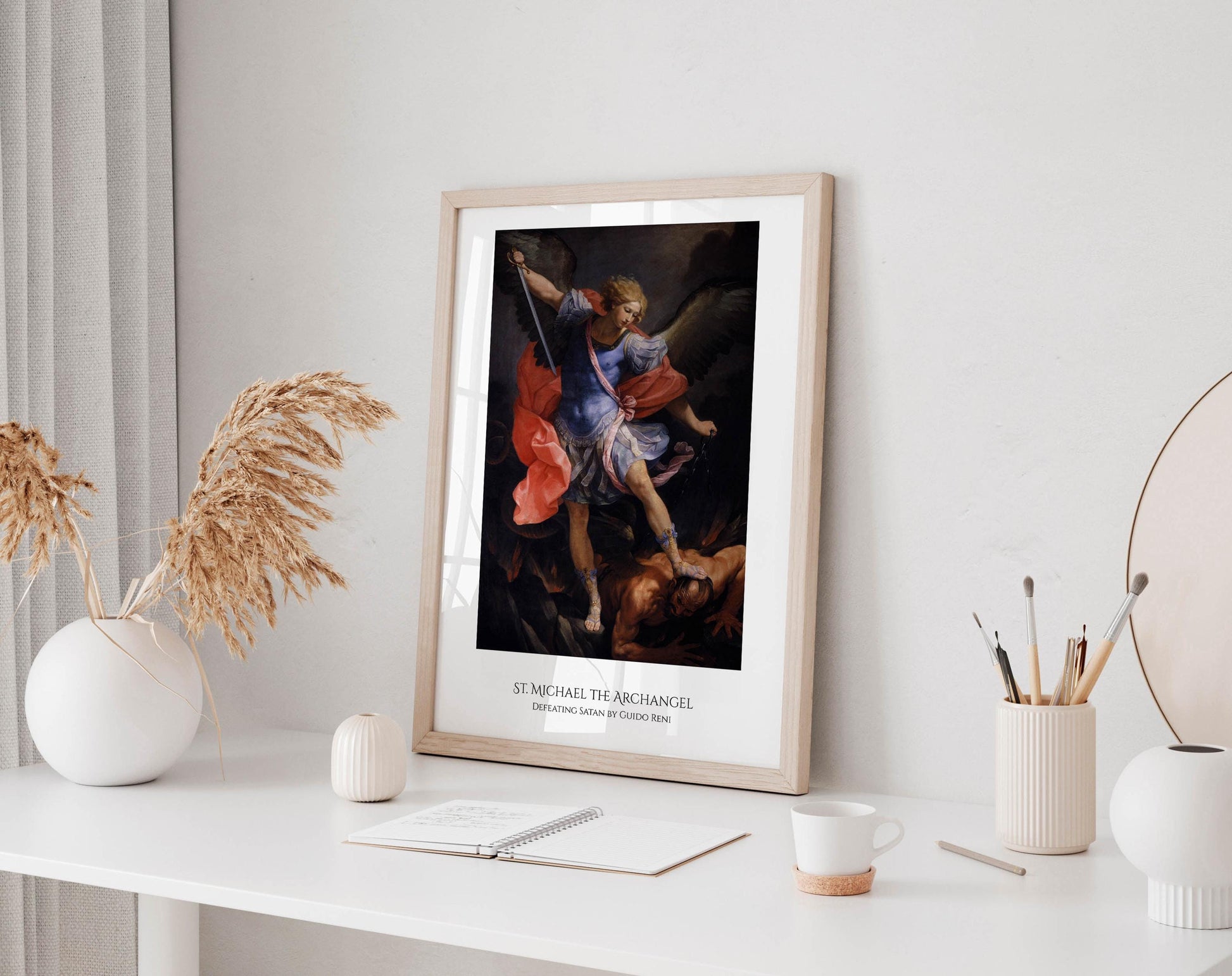 a framed painting of St. Michael, the archangel, with a white desk in the foreground. The painting depicts St. Michael holding a sword and a shield, with a figure in the background. The desk has various items on it, including a cup, a notebook, and a vase with dried flowers.