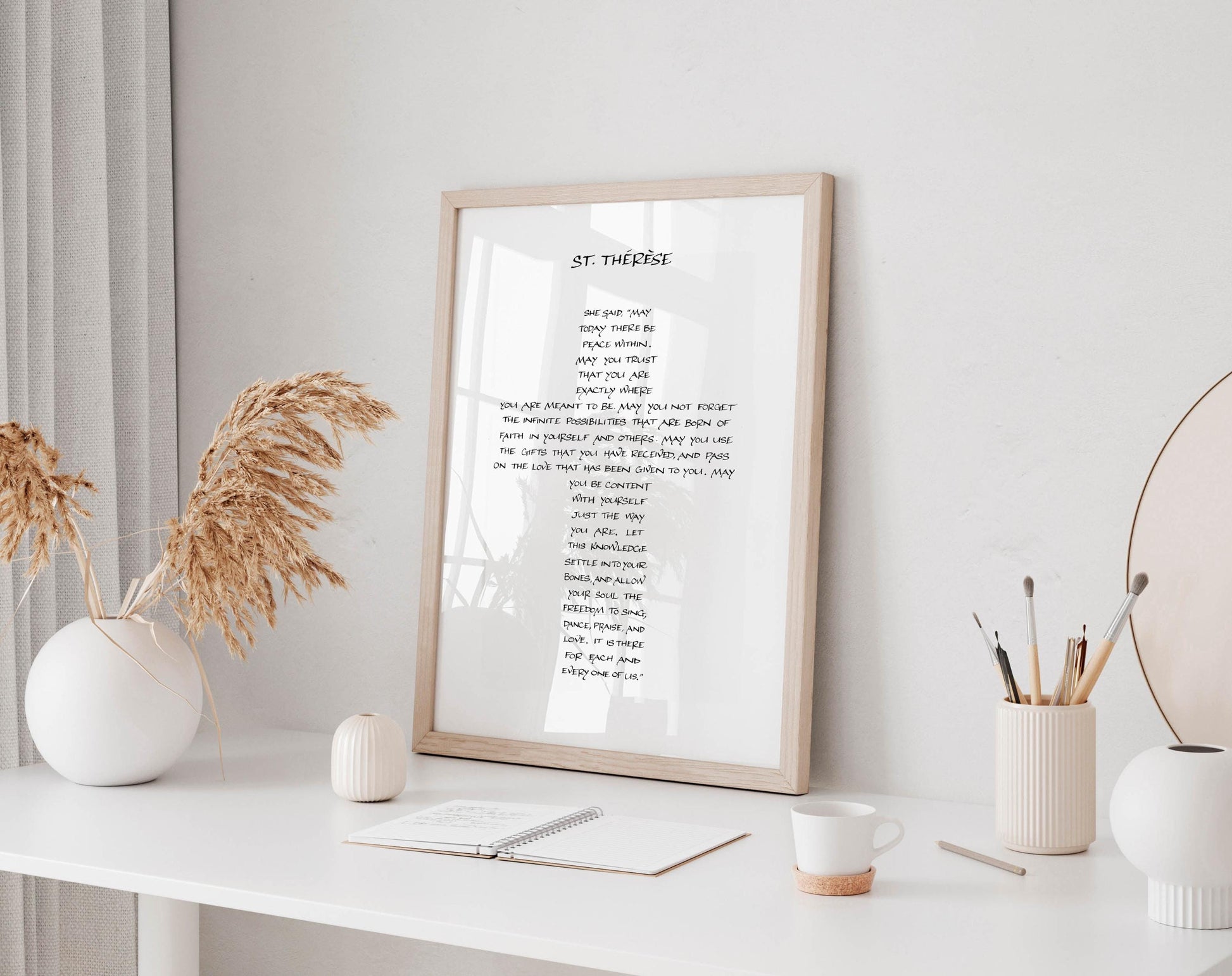 a framed poem titled "St. Therese" displayed on a white shelf, with a notebook, a cup, and a vase of dried flowers nearby.