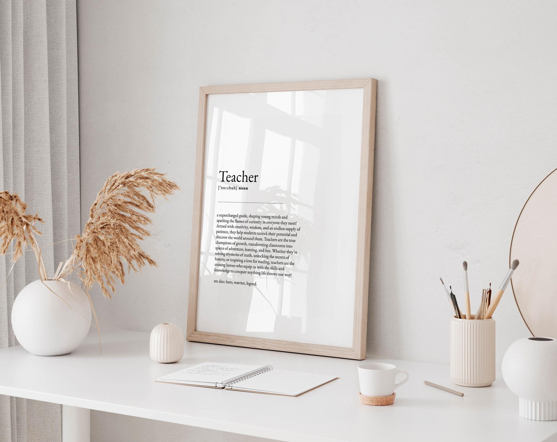 a desk with a framed definition of a teacher, a white vase with dried flowers, a white mug, a notebook, a pen holder, and a small white candle.