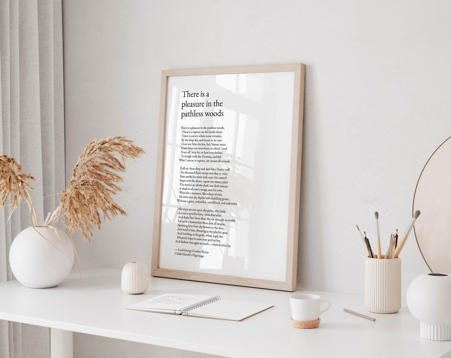 a framed poem titled "There is a pleasure in the pathless woods" displayed on a white desk, accompanied by a notebook, a cup, and a vase with dried flowers.