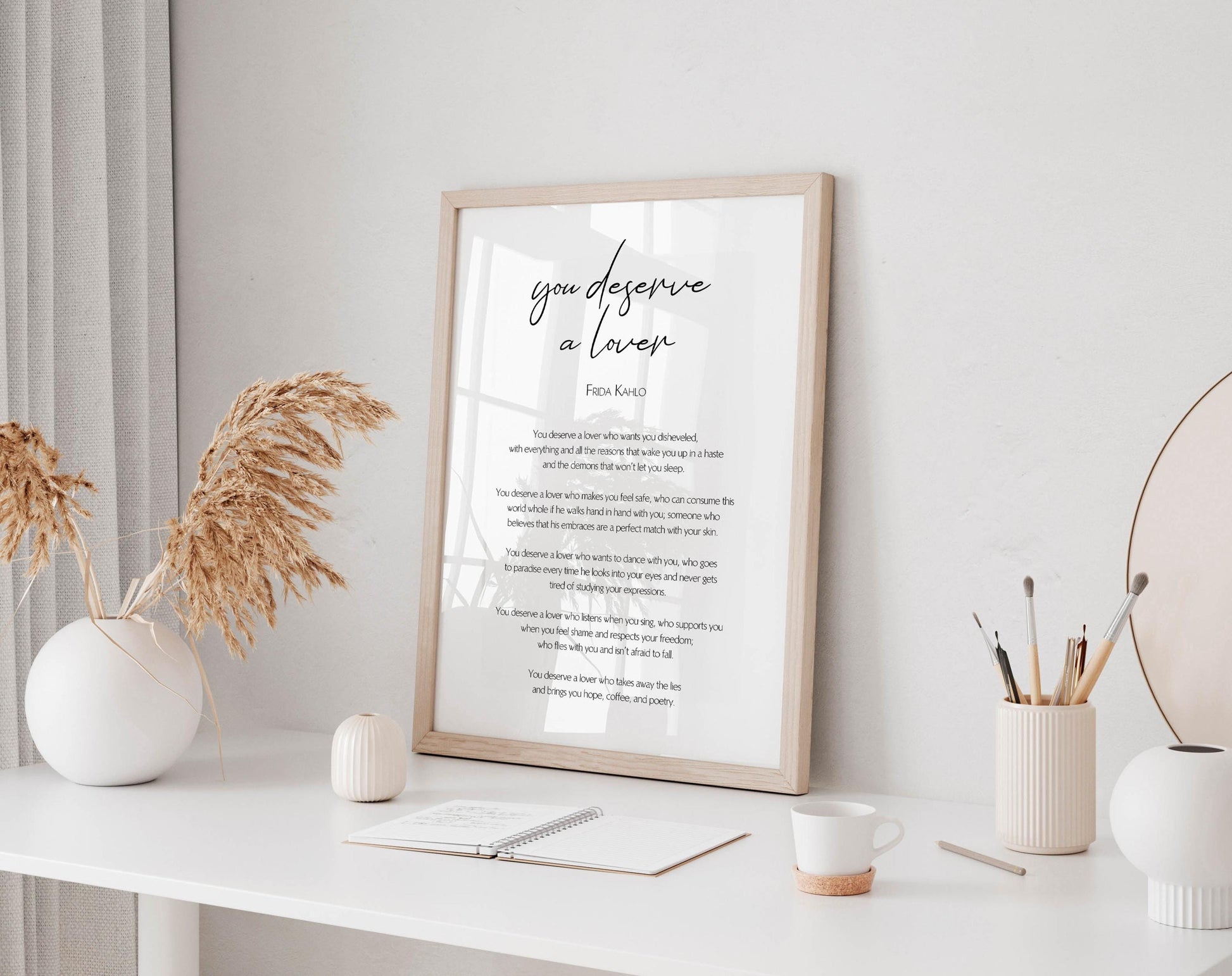 a framed poem titled "You deserve a love" displayed on a white desk, with a notebook, coffee cup, and decorative items such as a vase and a potted plant nearby.