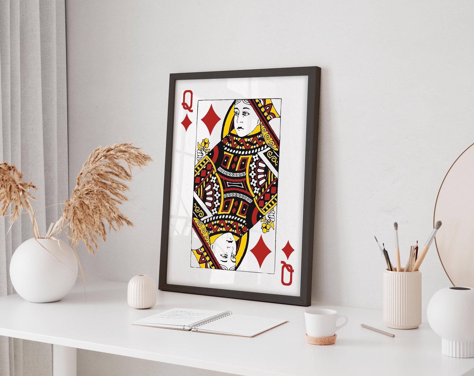 a framed illustration of a queen of spades playing card on a white desk, surrounded by various objects such as a vase, a cup, and a notebook.