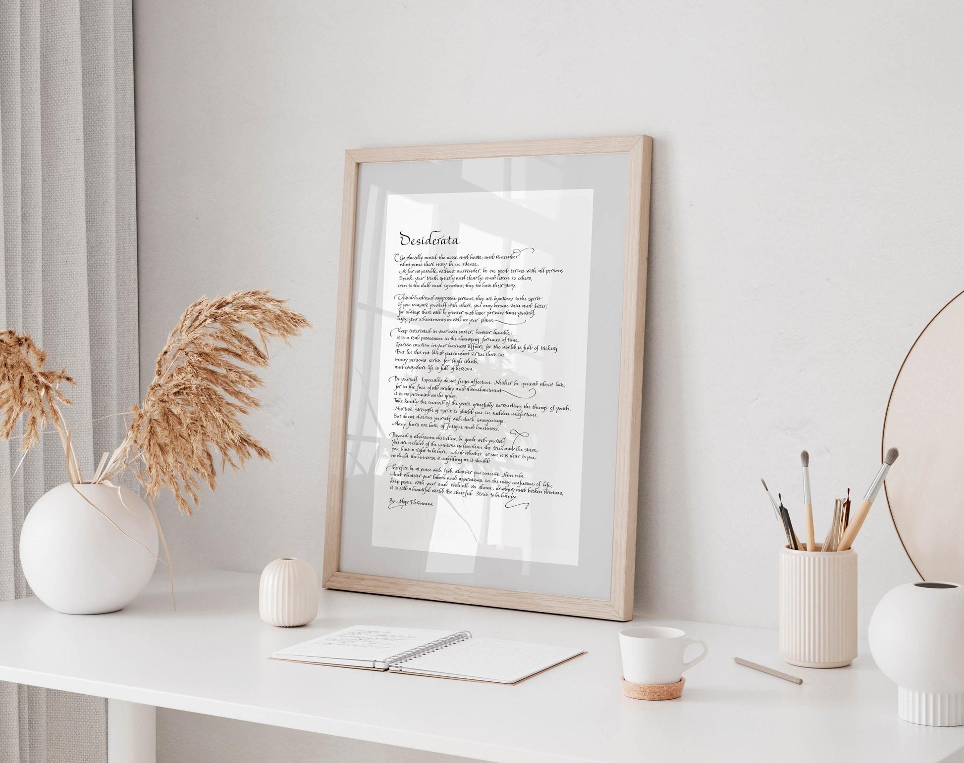 a framed text on a white desk, surrounded by various objects such as a cup, a notebook, and a vase with dried flowers.