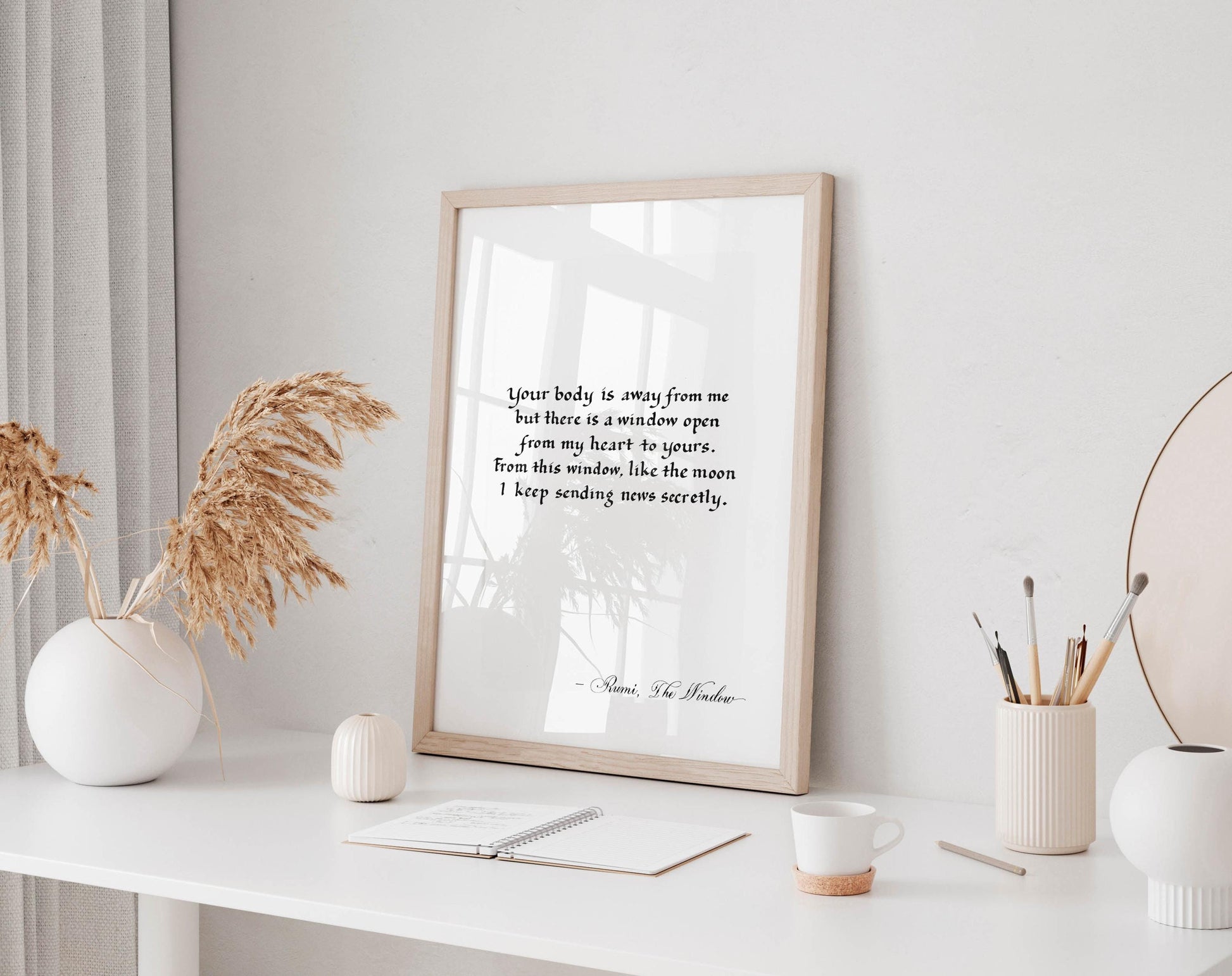 a white desk with a framed quote, a notebook, a cup, and a vase with dried flowers. The desk is set against a white wall, and there is a window in the background.