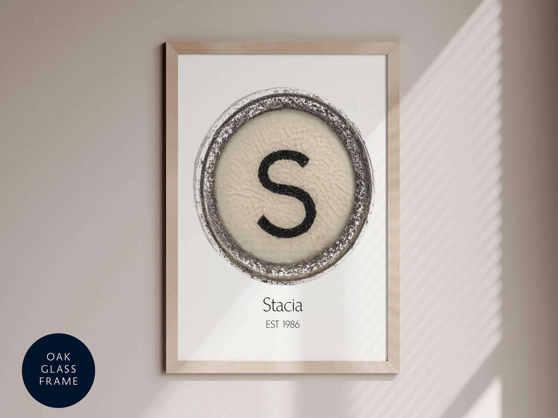 a framed print with a circular design containing a large letter 'S' in the center, surrounded by a decorative border. The print is titled "Stacia" and is framed in an oak-colored frame.